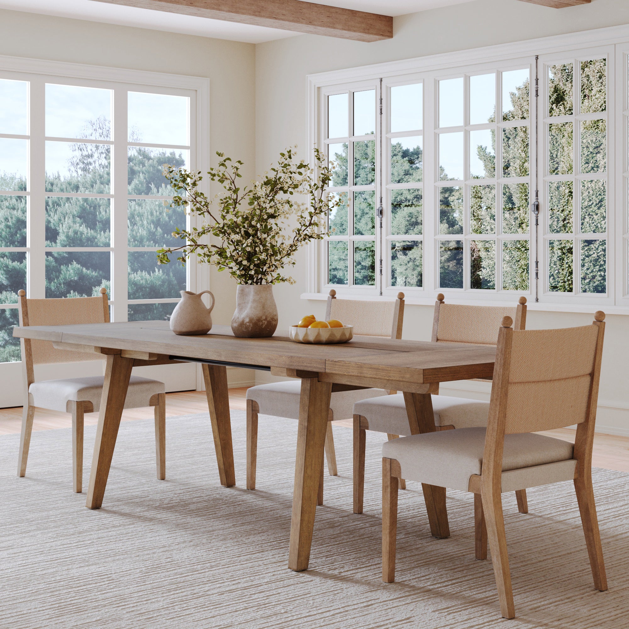 A bright dining room showcases the Nathan James Farmhouse Wooden Dining Table with Leaf Extension in light brown, four beige cushioned chairs, a matching rug, and a vase of green branches. Sunlight pours in, with views of trees outside.