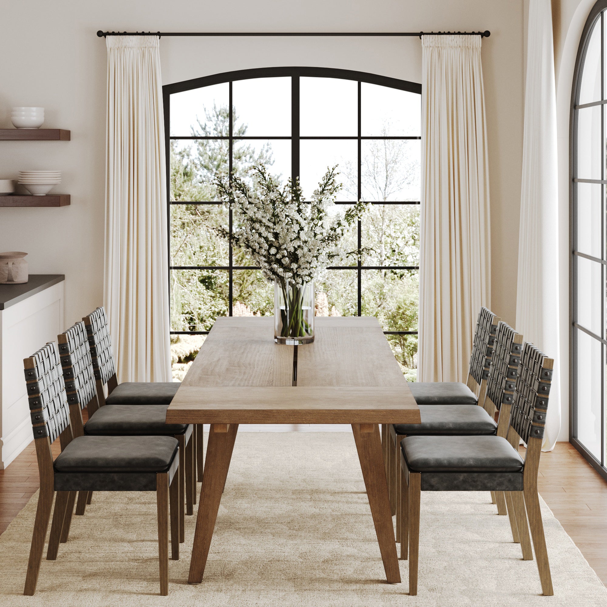 A modern dining room features the Nathan James Extendable Dining Set with 6 black dining chairs, a beige rug, and a vase of white flowers. Large windows with white curtains let in natural light and showcase greenery outside.