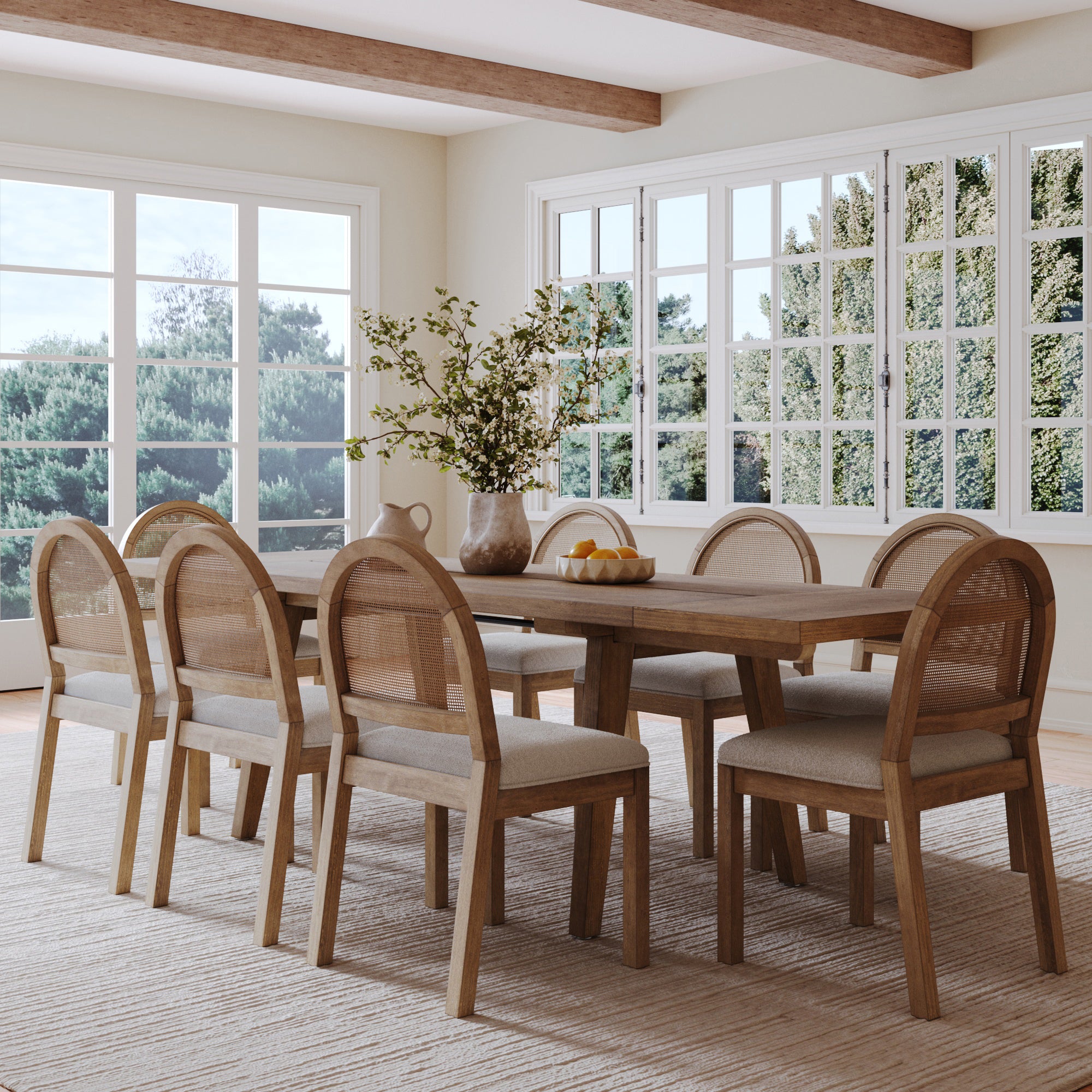 A bright dining room with large windows features the Nathan James Modern Farmhouse Table Extension with Oak Veneer Light Brown, solid wood legs, four upholstered chairs, a vase of greenery, and a bowl of lemons in a neutral décor filled with natural light.