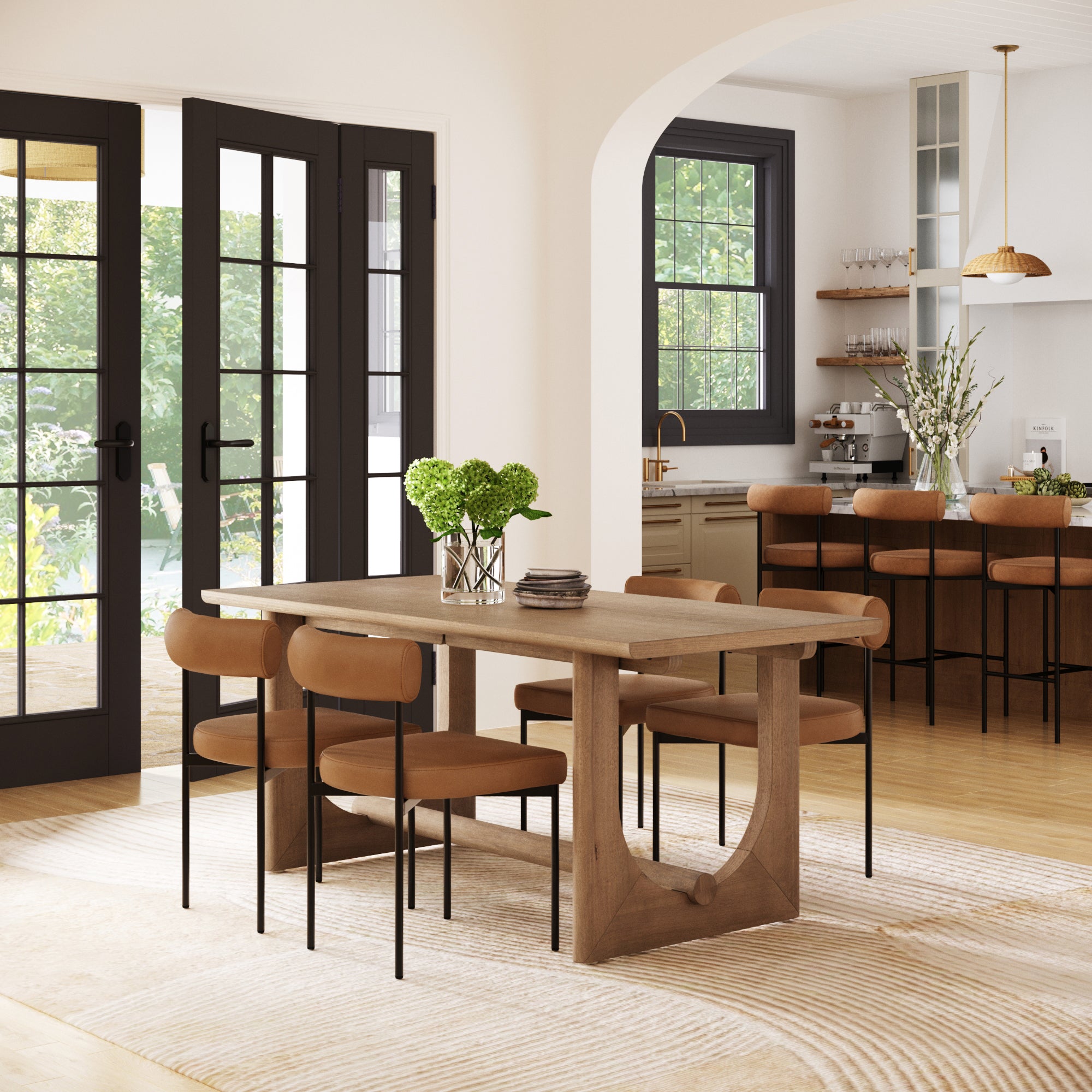 A modern dining room features the Nathan James Dining Room Set for 4 with faux leather chairs in light brown, a stylish table, green plant accents, large glass doors, an open kitchen with bar stools, and pendant lighting.