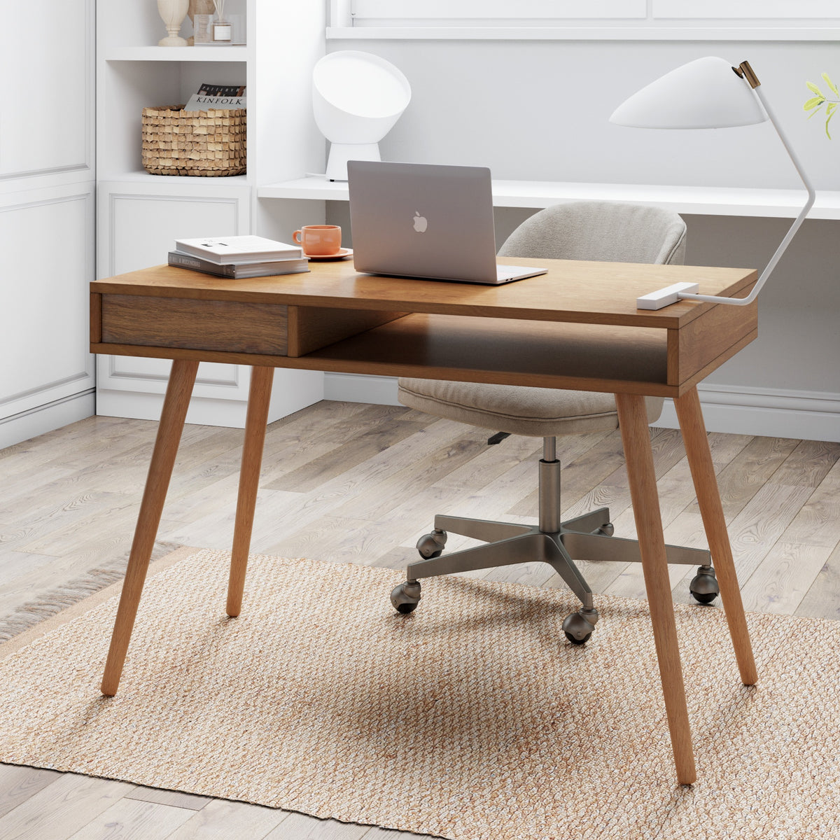 A wooden desk with angled legs holds a closed laptop, a mug, a notebook, and a desk lamp. A cushioned office chair is pushed in, and the room has light wood floors, a rug, and shelves in the background.