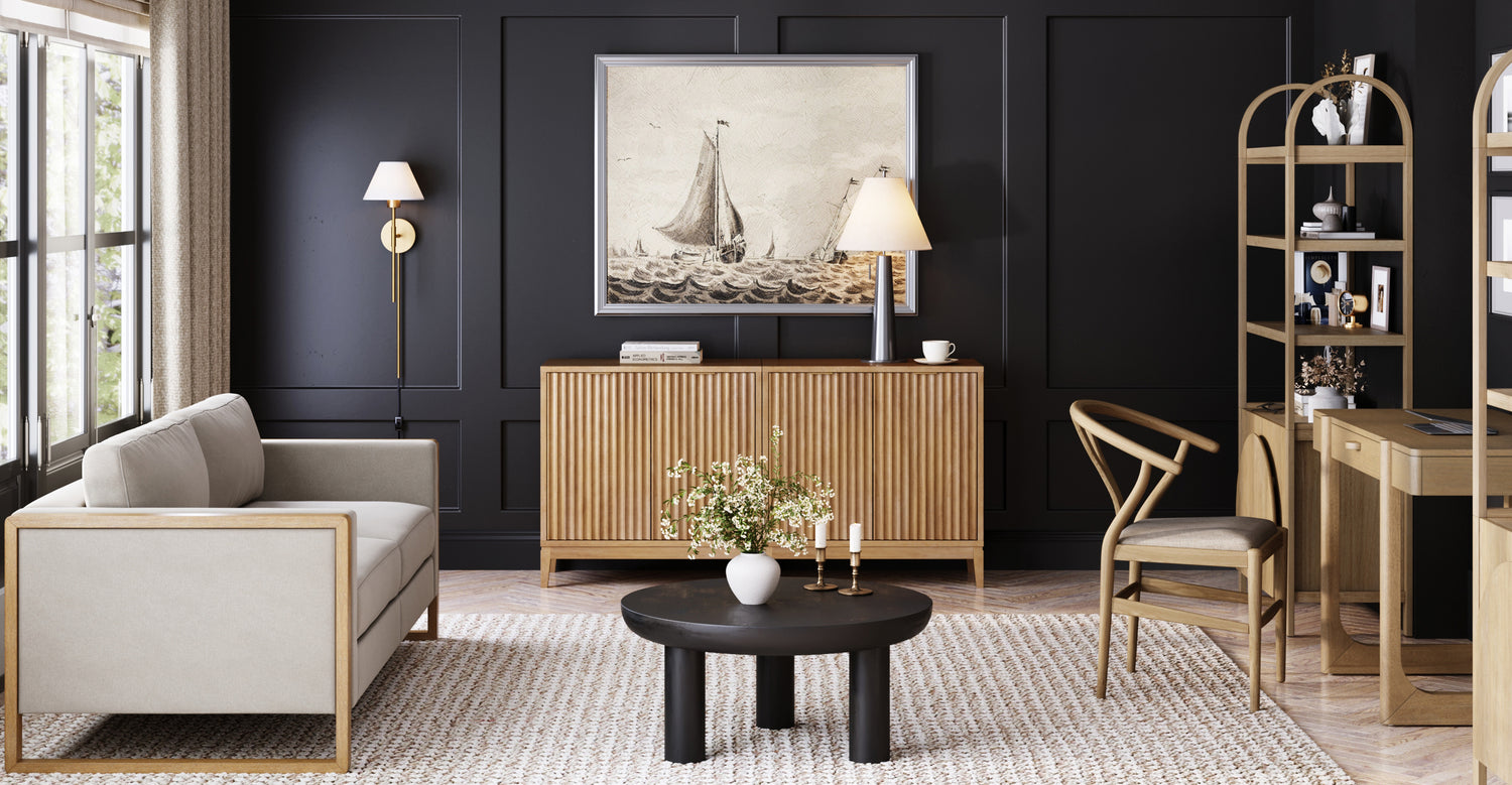 A modern living room with a light gray couch, wooden furniture, black paneled walls, a large window, and a framed sailboat artwork above a sideboard. A round black coffee table holds a vase with flowers and candles.