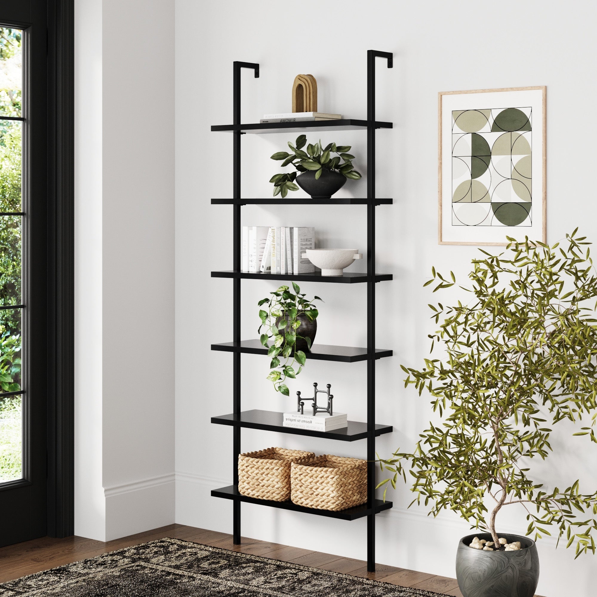 A modern black metal bookshelf with five shelves holds books, plants, baskets, and decorative items. A green leafy plant stands nearby, and abstract art hangs on the white wall. Sunlight enters through a glass door.