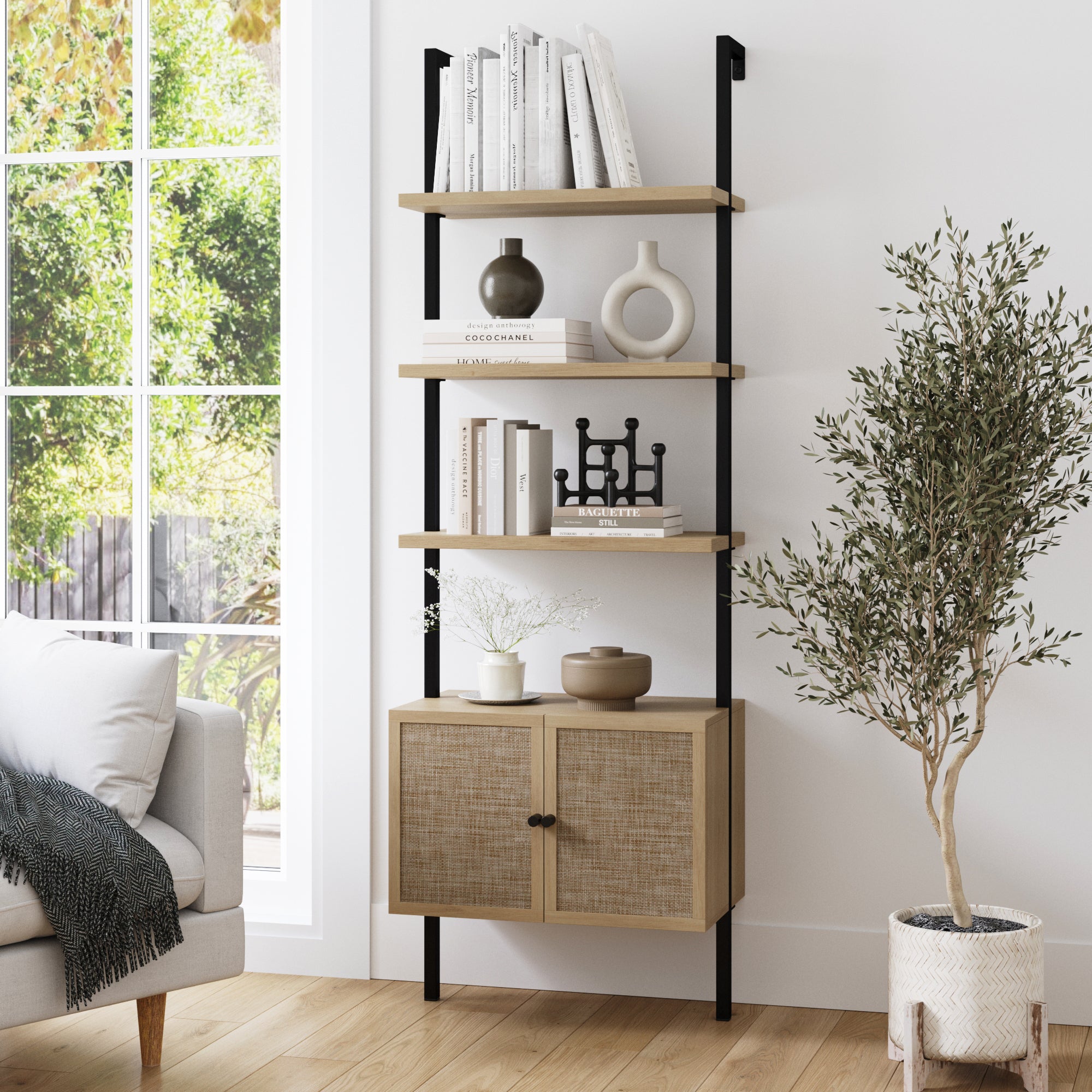 A modern wooden bookshelf with three open shelves displaying books, vases, and decor sits against a white wall next to a potted olive tree. Sunlight streams through a large window onto a light wood floor.