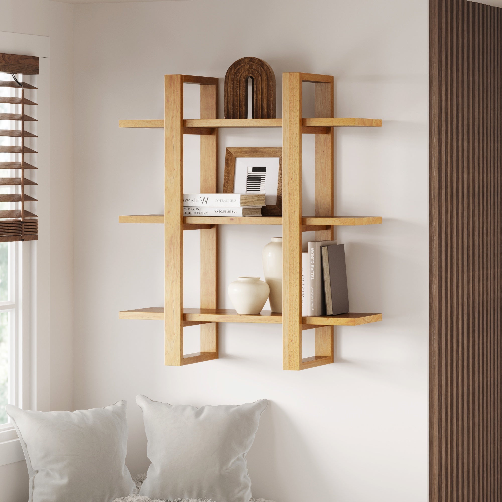 A modern wooden wall shelf holds decorative items, including two white vases, a black book, a framed photo, and abstract wooden art. The shelf is above a white cushioned seat near a window with blinds.