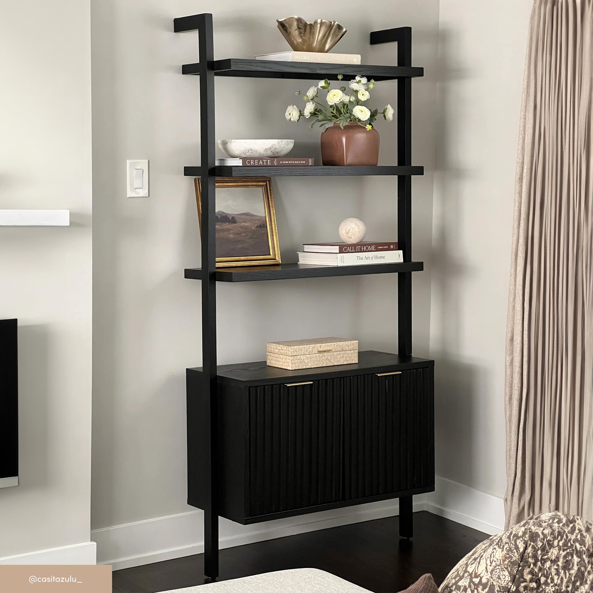 A modern black shelving unit with three open shelves and a cabinet at the bottom, displaying books, decorative vases, a gold bowl, and a framed landscape photo against a light-colored wall.