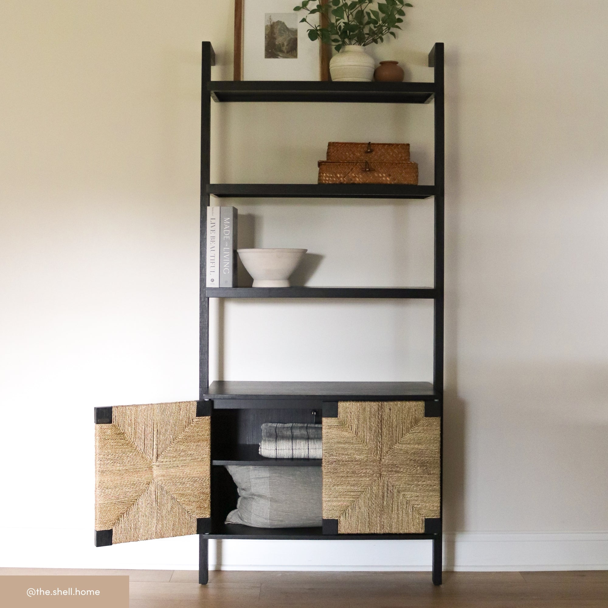 A black shelving unit with open shelves and two lower woven cabinet doors stands against a light wall. Shelves display a plant, baskets, books, a framed picture, and a white bowl.