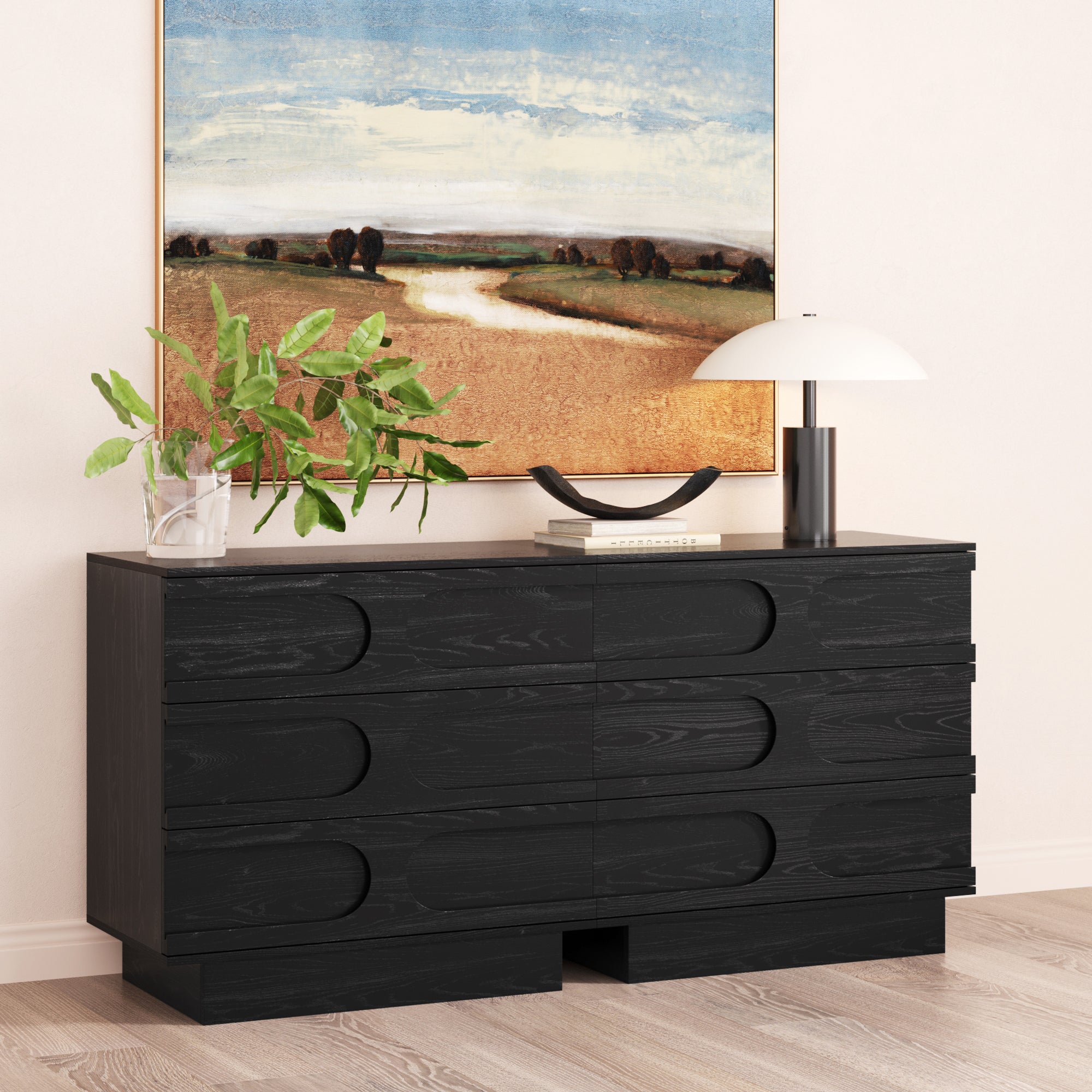 The Nathan James Mid-Century 3-Drawer Arched Dresser in Black Oak (Set of 2) features rounded handles and sits against a light wall, topped with a potted plant, books, lamp, and decor. A landscape painting hangs above this stylish scene.