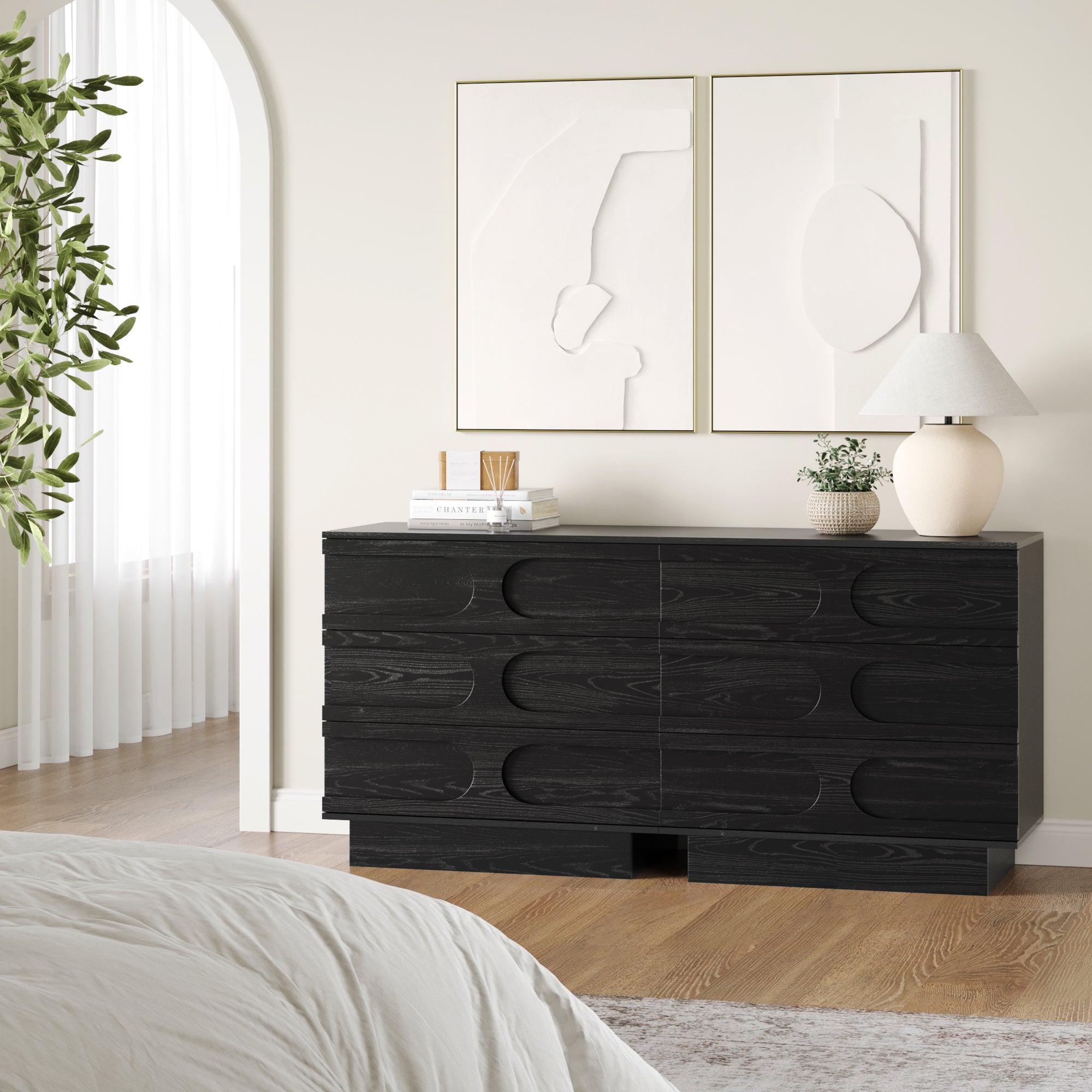 A modern bedroom showcases two Nathan James Mid-Century 3-Drawer Arched Dressers in Black Oak, topped with a lamp, plant, and decor, flanked by abstract art above and a leafy plant near sheer-curtained windows for a stylish mid-century vibe.