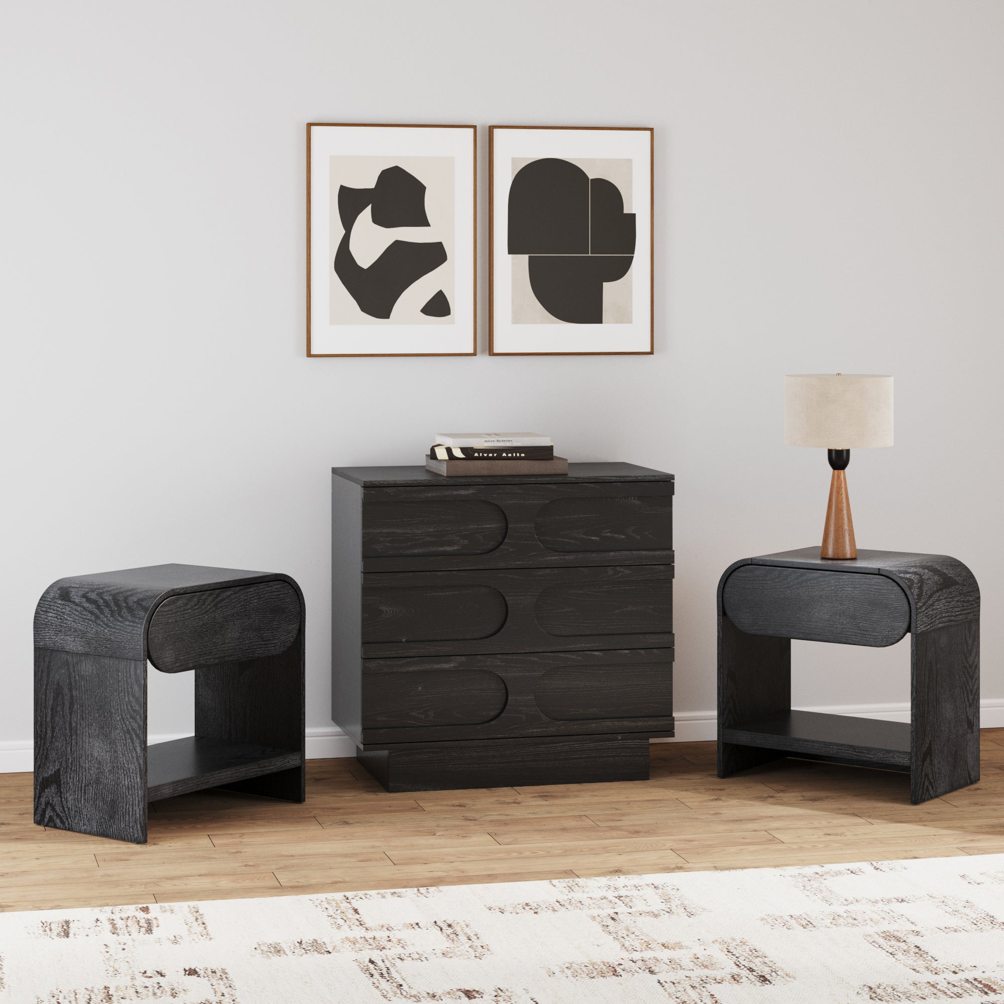 A modern bedroom features the Nathan James Mid-Century Arched Dresser and Two Nightstands Set in black oak, a table lamp, stacked books, two abstract black-and-white artworks on a white wall, wood floors, and a patterned rug.