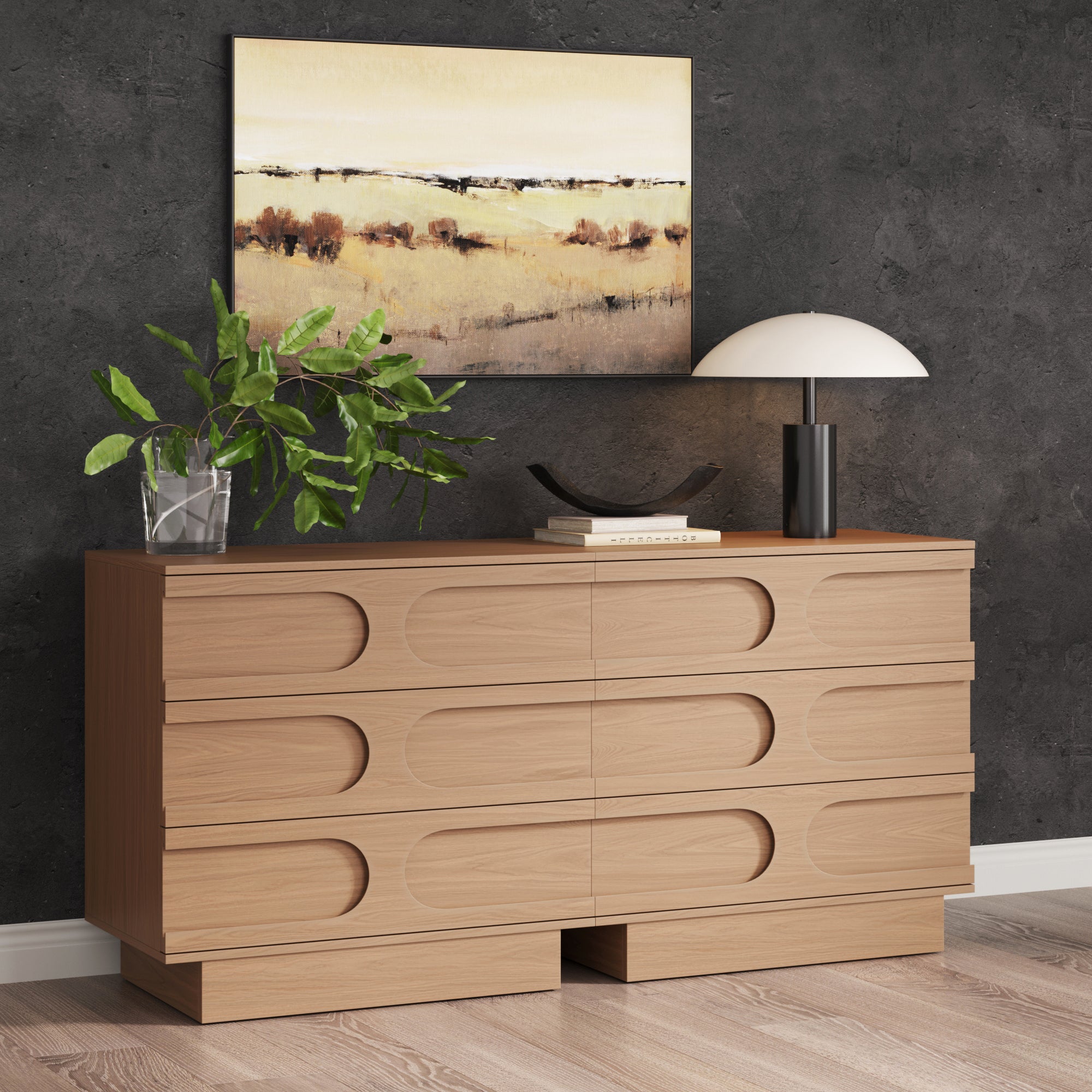 The Nathan James Mid-Century 3-Drawer Arched Dresser in Raw Oak (Set of 2) stands against a dark textured wall, topped with a plant in a glass vase, books, and a modern lamp. A landscape painting above completes the elegant mid-century look.