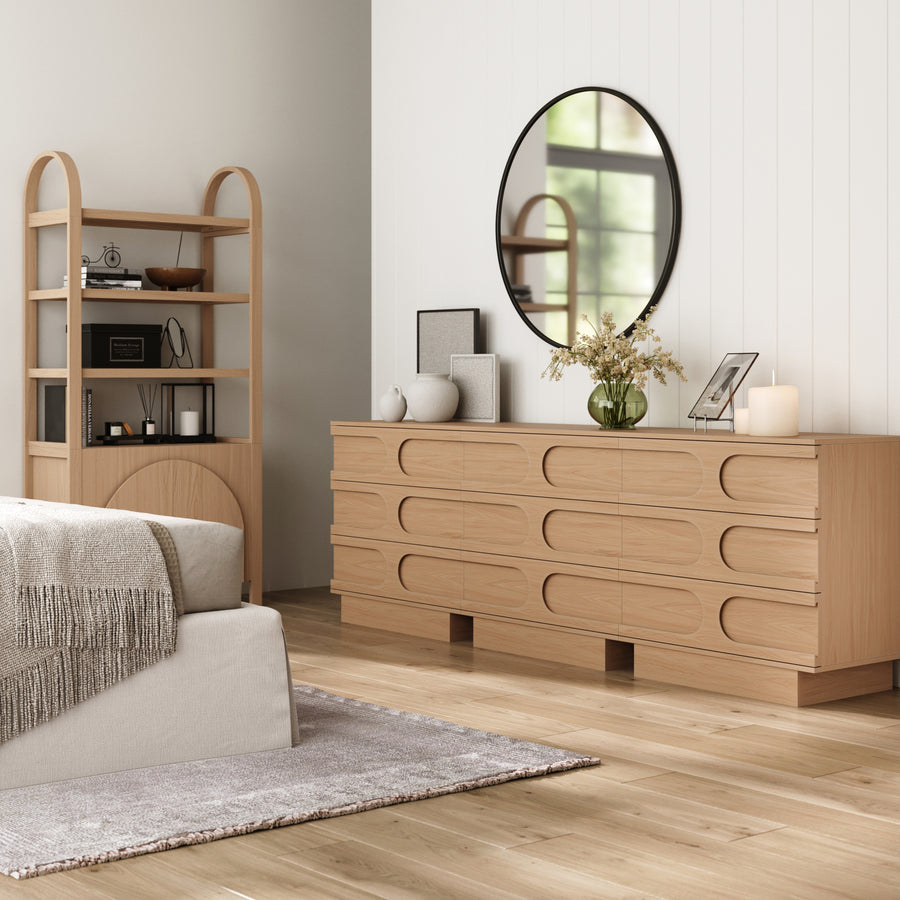 A modern bedroom with light wood floors, a beige bed and gray throw, the Nathan James Mid-Century 3-Drawer Arched Dresser Raw Oak (Set of 3), a round mirror, vase of flowers, decor accents, and a wooden shelf filled with books and accessories.
