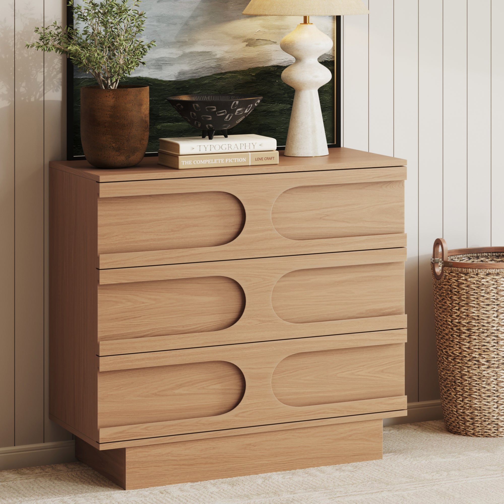 The Nathan James Mid-Century 3-Drawer Arched Dresser in black oak showcases a curved silhouette and three drawers. Styled against a beige paneled wall, it’s topped with books, décor, and a woven basket nearby for a modern look.
