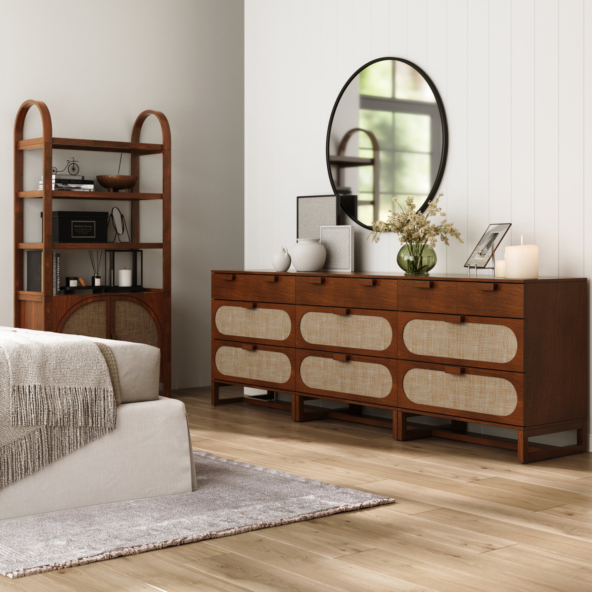 A modern living room with a light wood floor, beige sofa with gray throw, Nathan James Mid-Century Rattan 3-Drawer Dressers in Dark Acacia (Set of 3), a round mirror above, vase of flowers, and shelves displaying books and decor.
