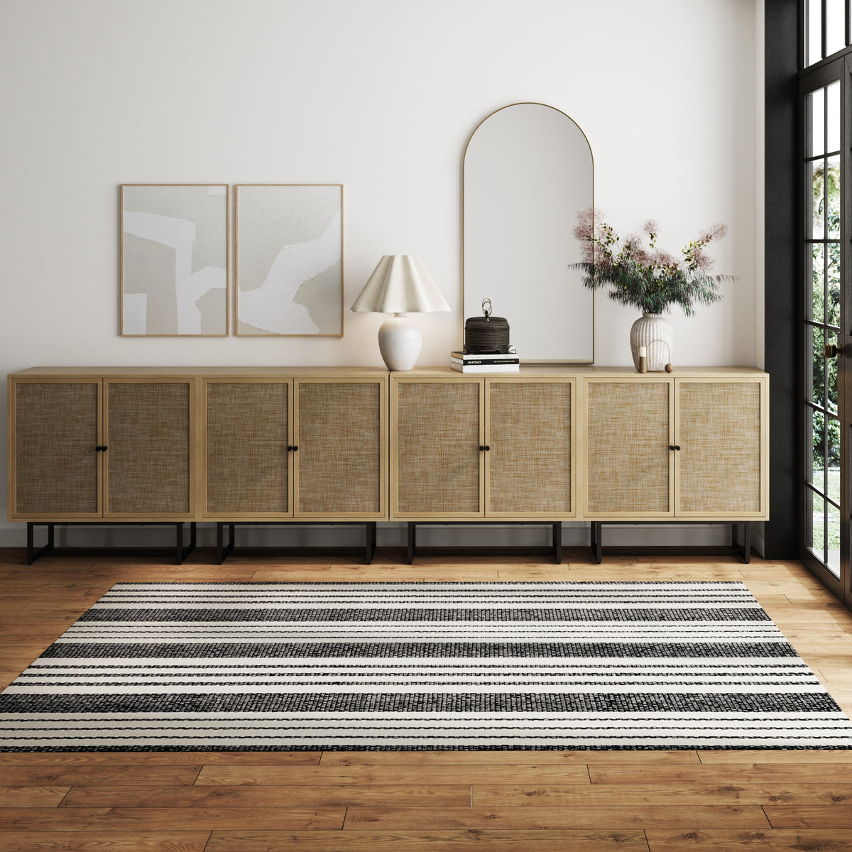 A modern minimalist room with a long wooden sideboard, a striped black-and-white rug, abstract wall art, a lamp, a mirror, and a vase of flowers, all set against a white wall and large windows.