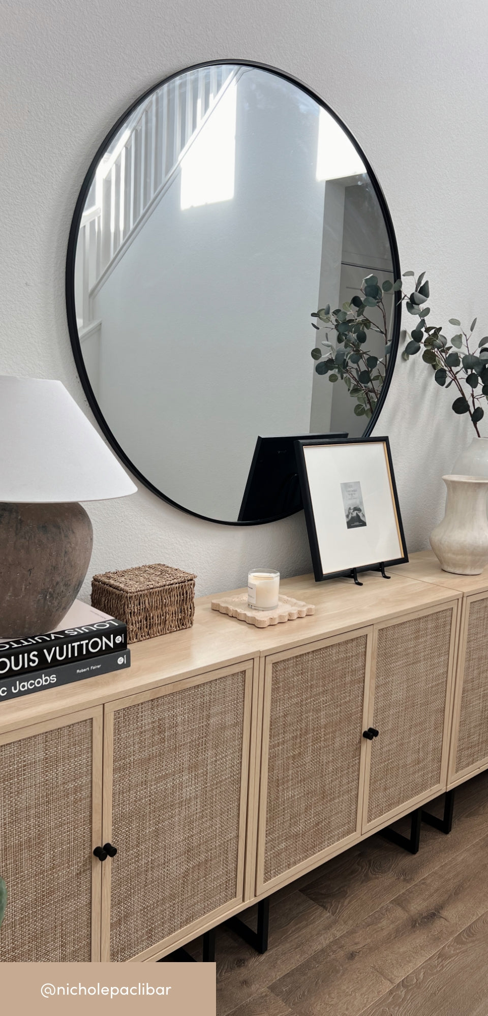 A round black-framed mirror hangs above a light wood console with rattan doors. The console is decorated with stacked books, a lamp, a candle, a small framed photo, and vases with greenery. Stairs are reflected in the mirror.
