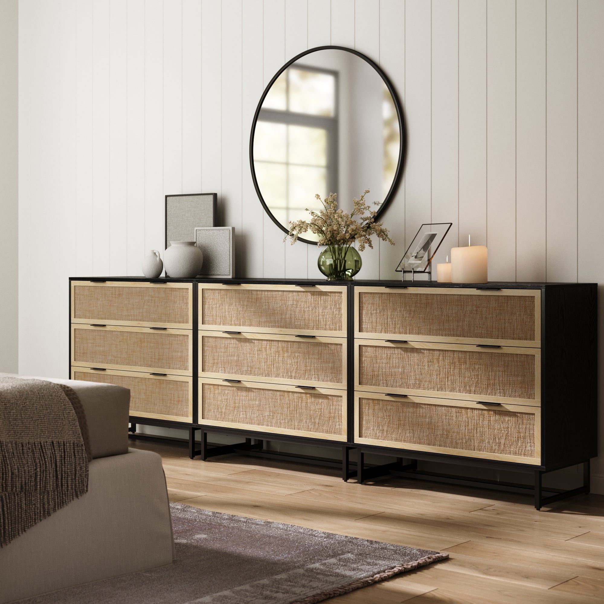 The Nathan James Rattan, Wood & Metal 3-Drawer Dressers in black (set of 3) stand against a white paneled wall near the bed and gray rug, topped with a round mirror, candles, framed photos, and a flower-filled vase.