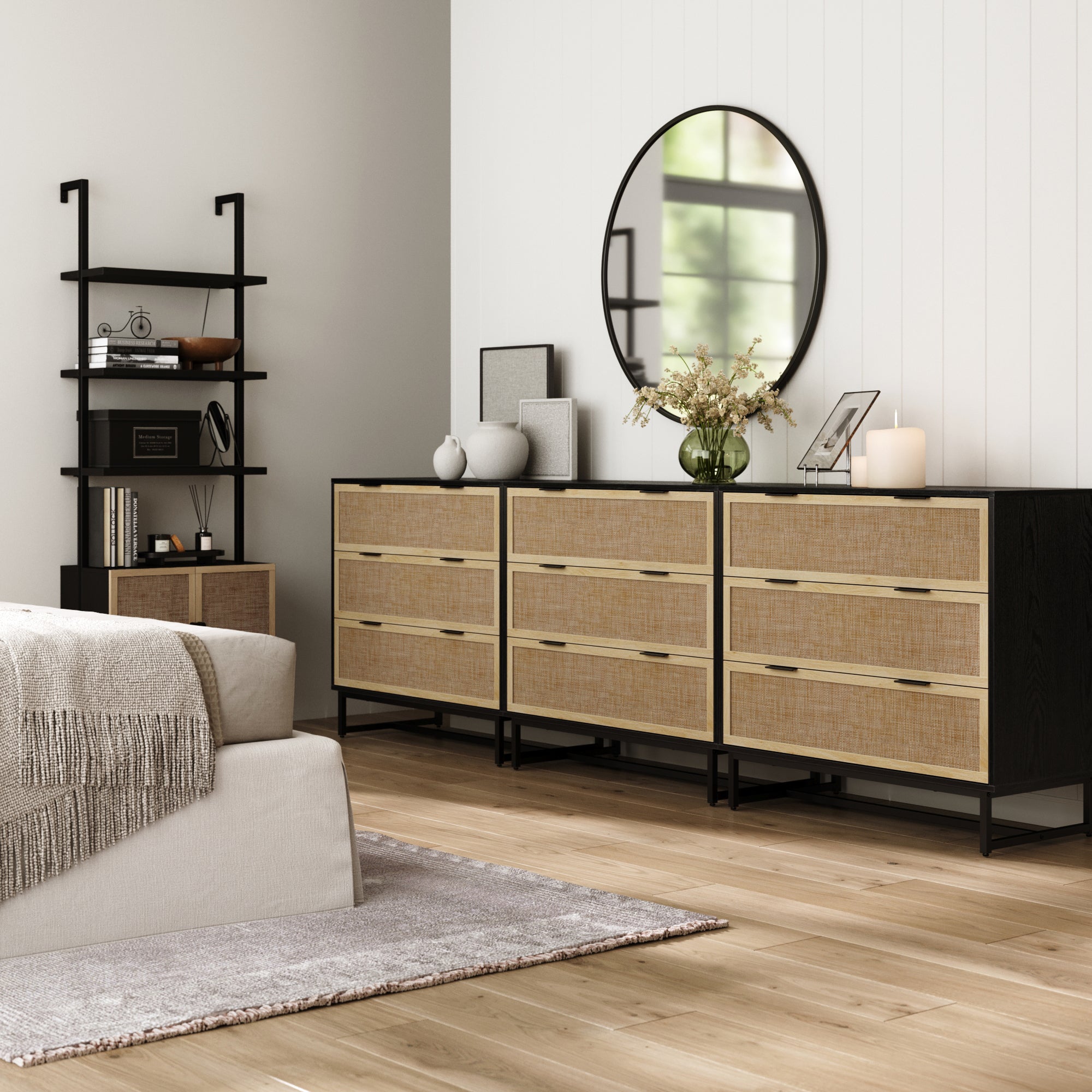 A modern bedroom features a bed with a gray blanket, three black Nathan James Rattan, Wood & Metal 3-Drawer Dressers, a round mirror above, a small shelf with books and decor, and light wood flooring.