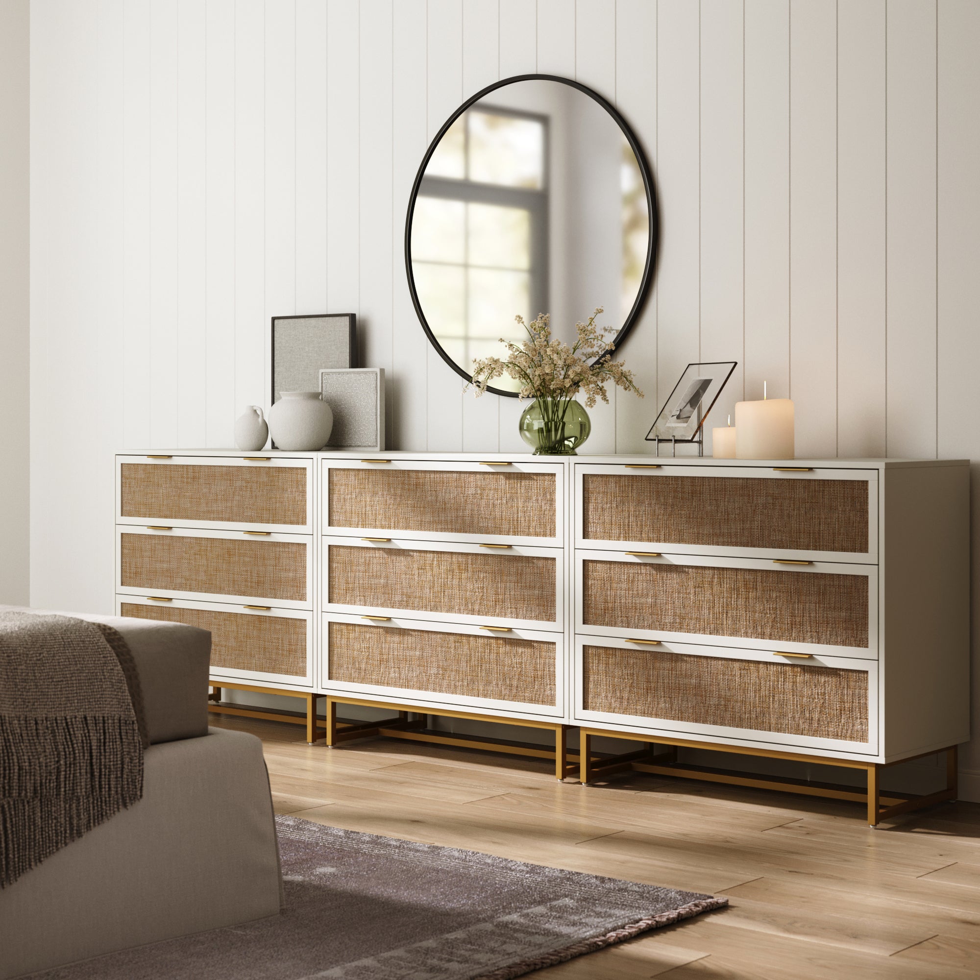 A modern bedroom showcases the Nathan James Rattan, Wood & Metal 3-Drawer Dressers White (Set of 3) with woven fronts, topped with a round mirror, vases, books, a candle, and flowers. Light wood floors and neutral decor complete the cozy vibe.