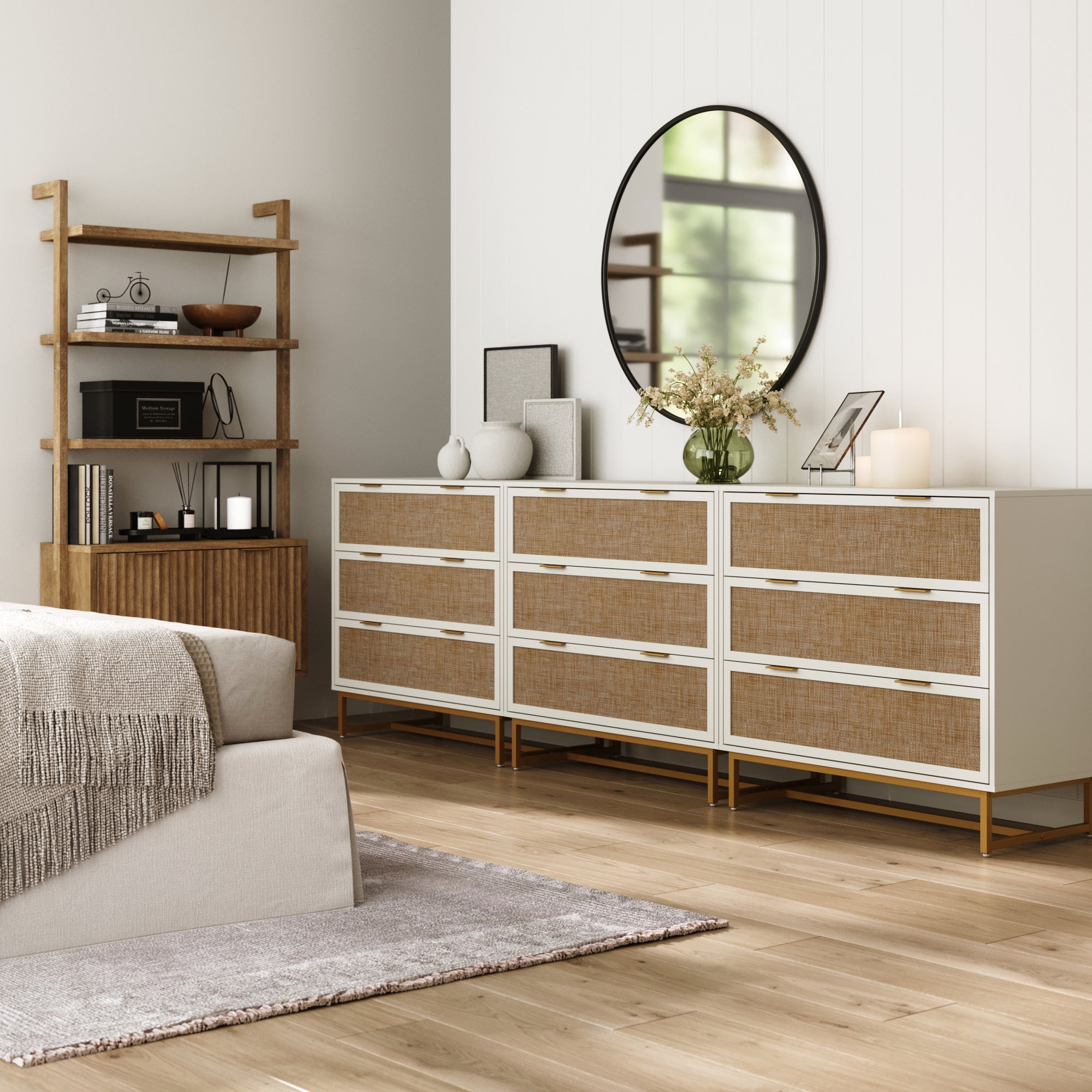 A modern bedroom with light wood floors, a beige bed, gray rug, white paneled walls, shelves holding decor and plants, and a set of three Nathan James Rattan, Wood & Metal 3-Drawer Dressers in white with a large round mirror above.