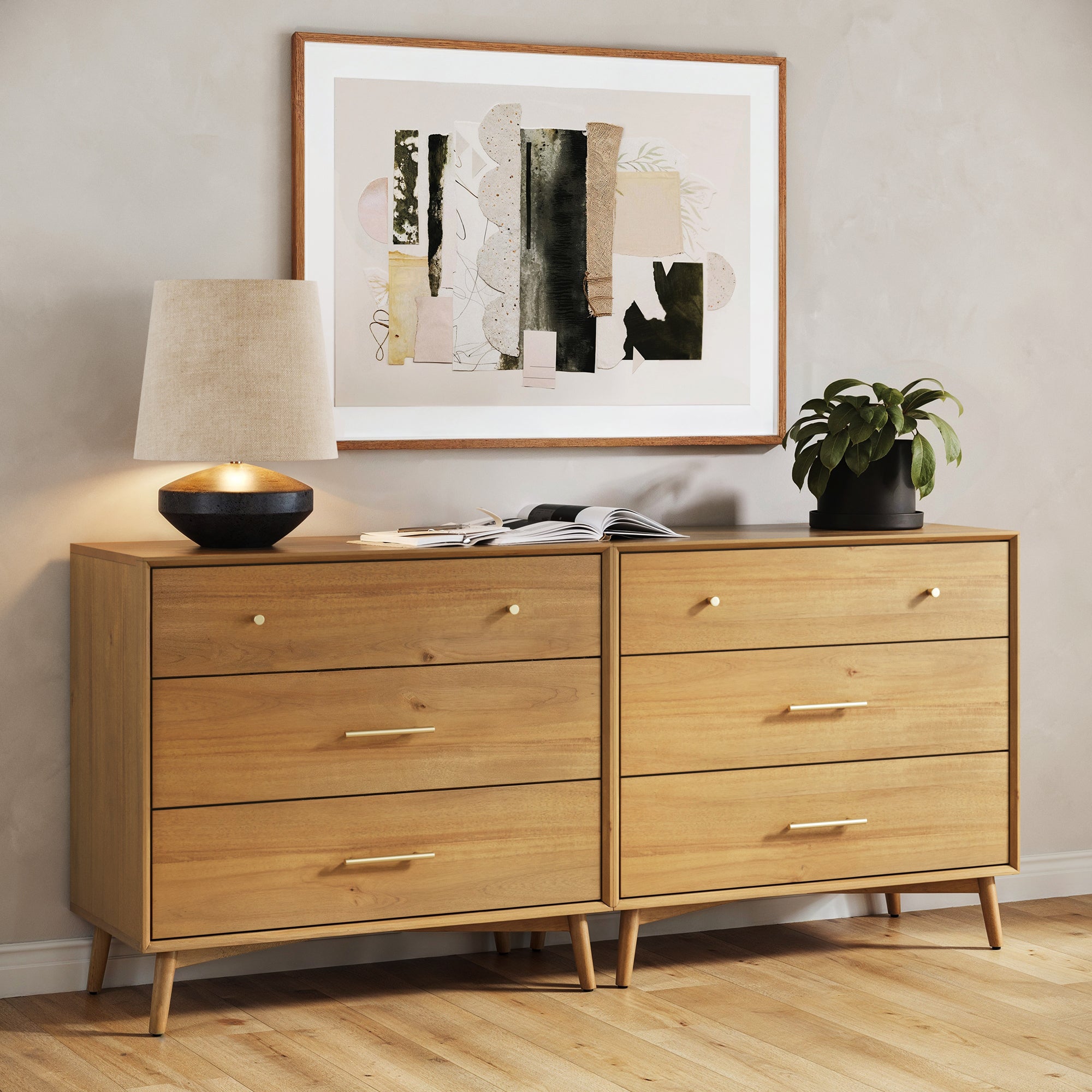 The Nathan James Mid-Century Modern Acacia 3 Drawer Dresser Set of 2 stands against a light wall, topped with a lamp, an open book, and a potted plant. Above the wood veneer dressers hangs abstract framed artwork.