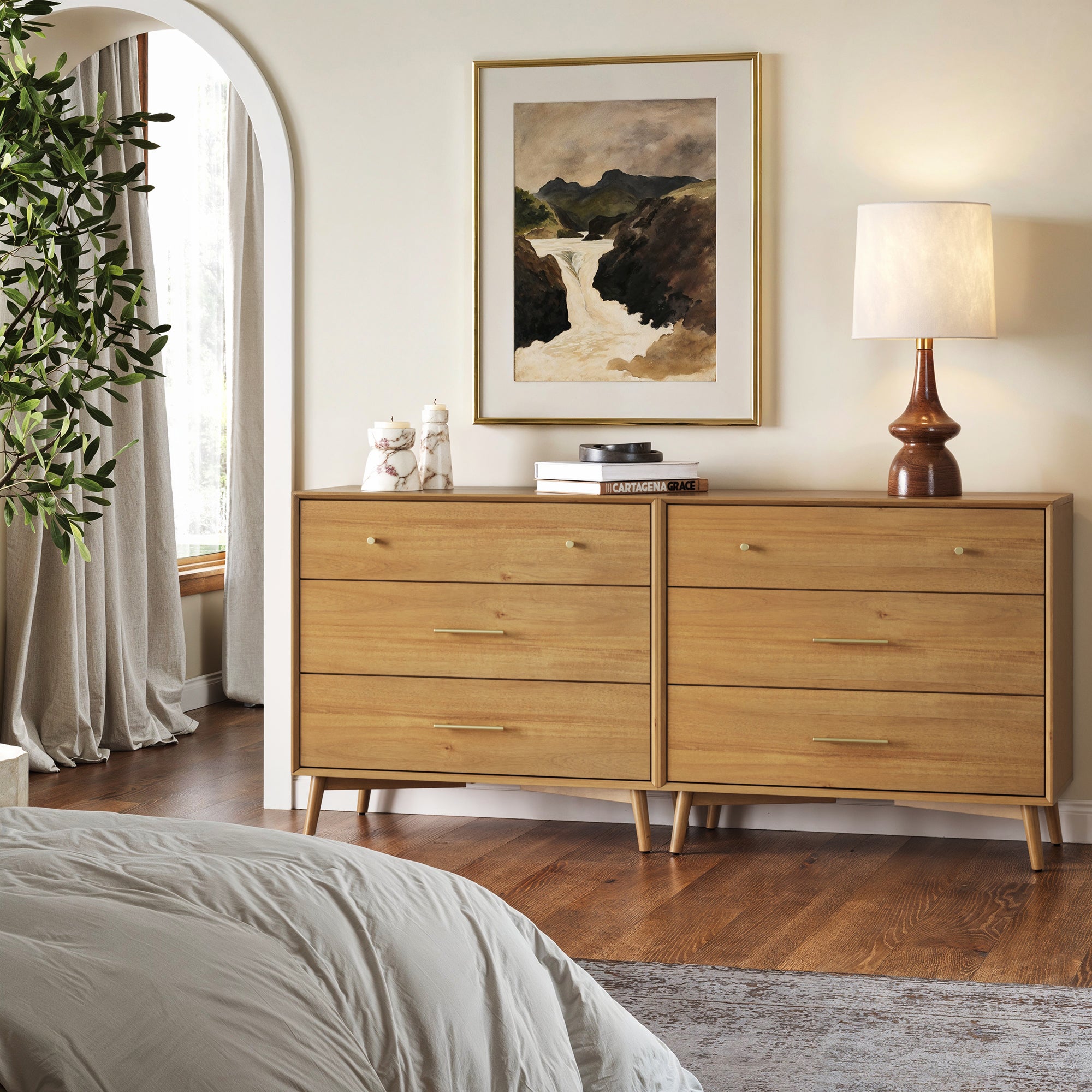 A bedroom features two Nathan James Mid-Century Modern Acacia 3 Drawer Dressers, topped with books, a white ceramic decor, and a lamp. Above them hangs a framed landscape painting, with part of a bed and large leafy plant visible.