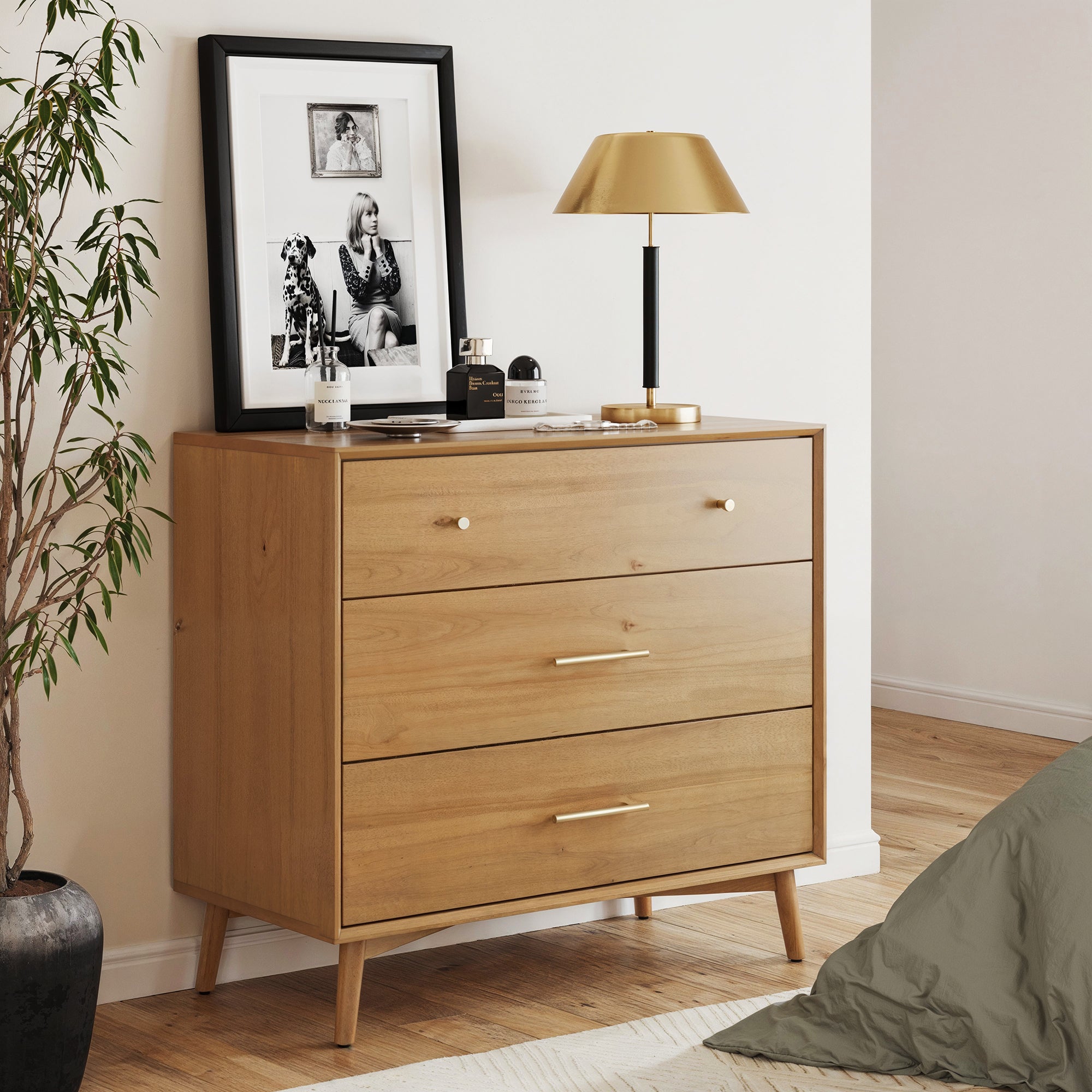 The Nathan James Mid-Century Modern Acacia 3 Drawer Dresser displays a gold lamp, framed black-and-white photo, perfume bottles, and a book. Nearby sits a potted plant and a green cushion on the wooden floor.