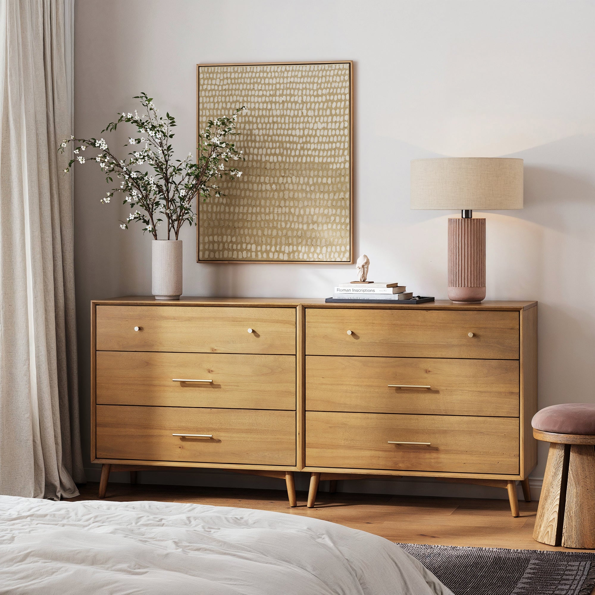 A modern bedroom showcases the Nathan James Mid-Century Modern Acacia 3 Drawer Dresser with gold handles, adorned with a lamp, books, a small sculpture, and a vase. Textured art hangs above; curtains and part of the bed are also visible.