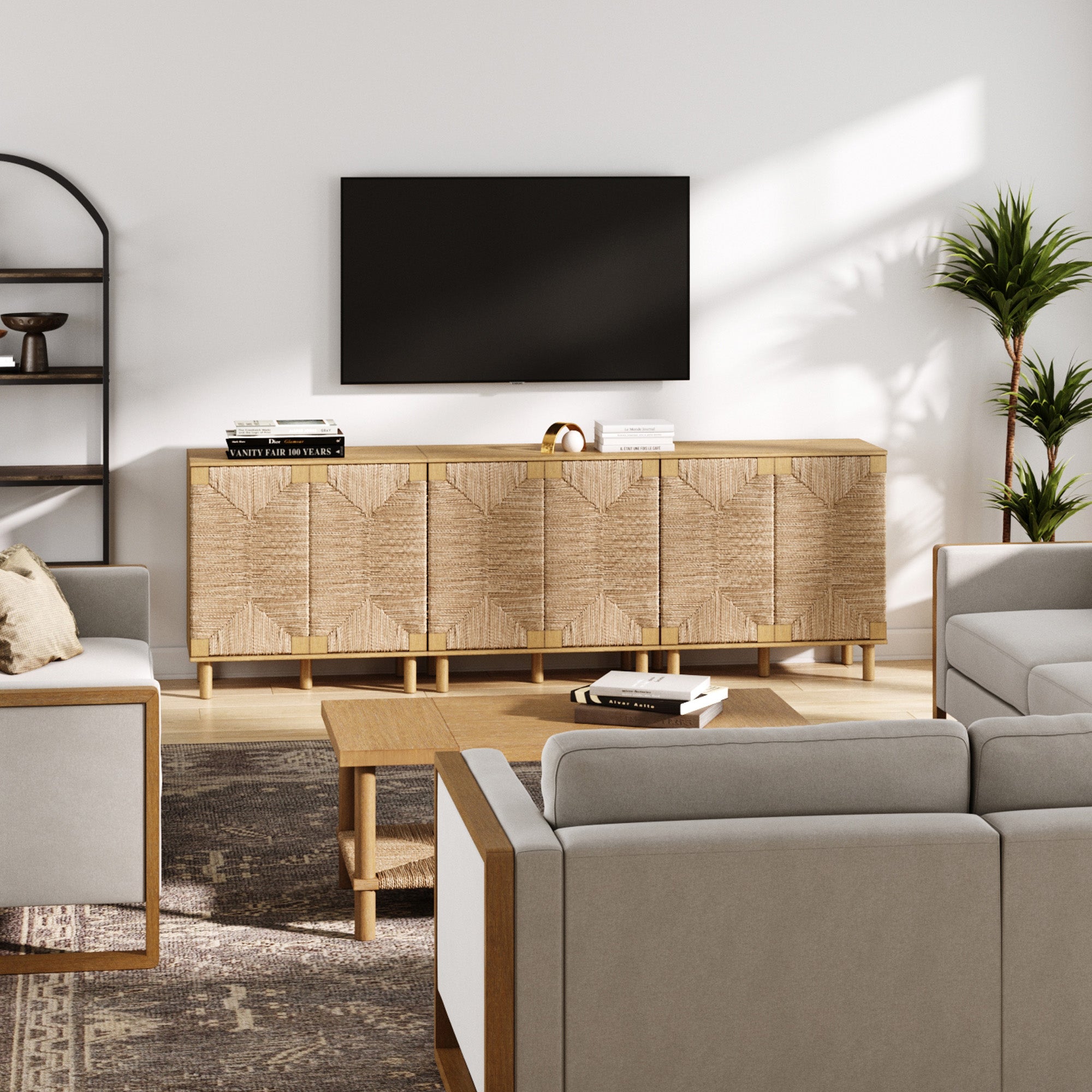 A modern living room with neutral tones, featuring a woven wood sideboard, wall-mounted TV, beige sofas, a wooden coffee table, indoor plants, and a black metal shelf against a white wall. Sunlight streams in from the right.