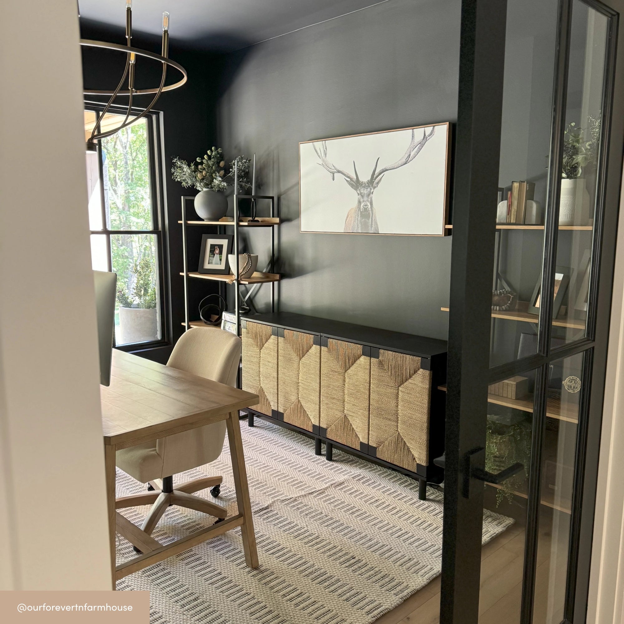 A modern home office with dark walls, light wood desk and chair, geometric rug, open shelving with decor, and a sideboard. A framed deer artwork hangs above the sideboard. Large windows let in natural light.