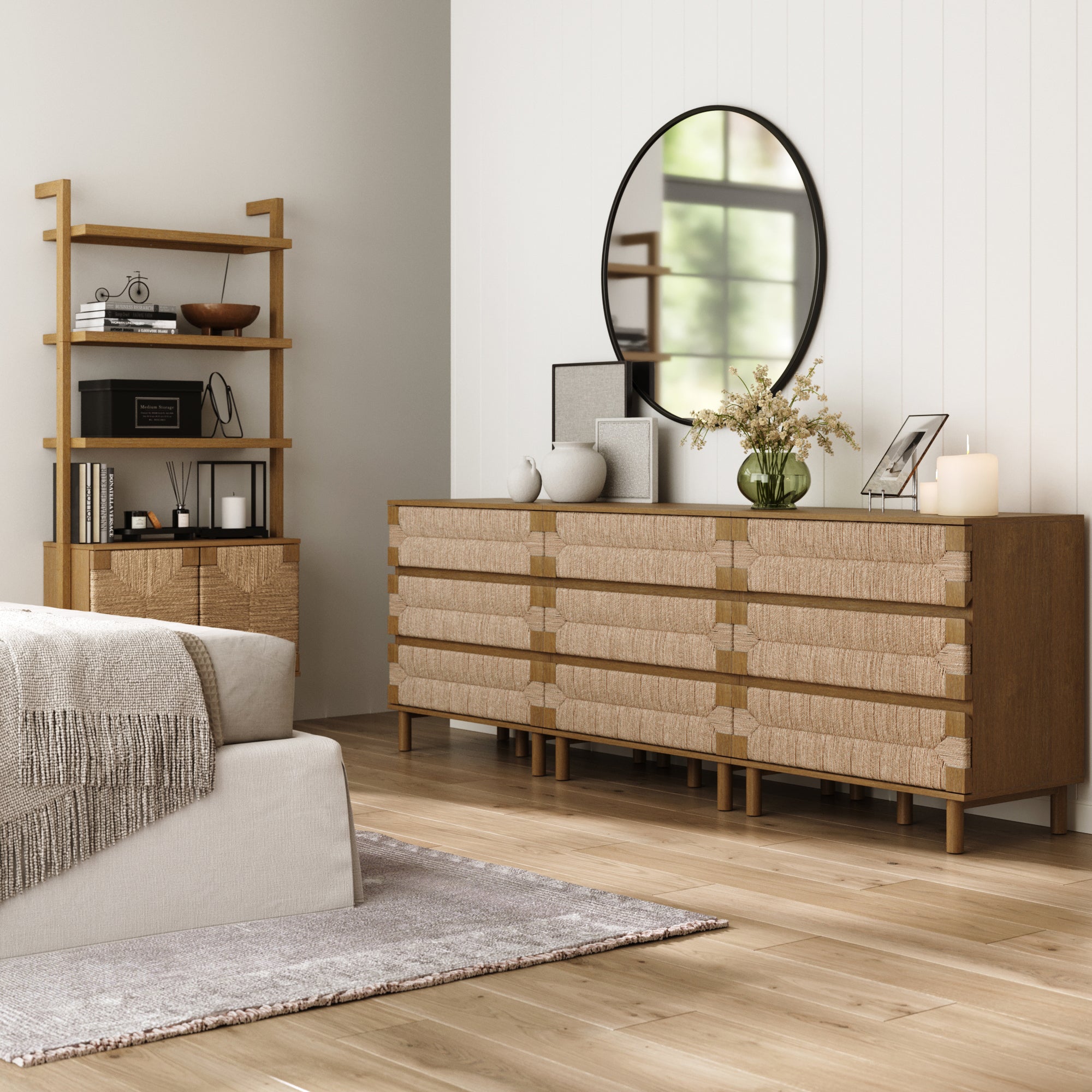 A modern bedroom styled with the Nathan James Boho Seagrass 3-Drawer Dresser Set in light brown, an oval mirror, decorative vases, a lit candle, a wooden shelf with books and decor, light walls, wood flooring, and a cozy bed.