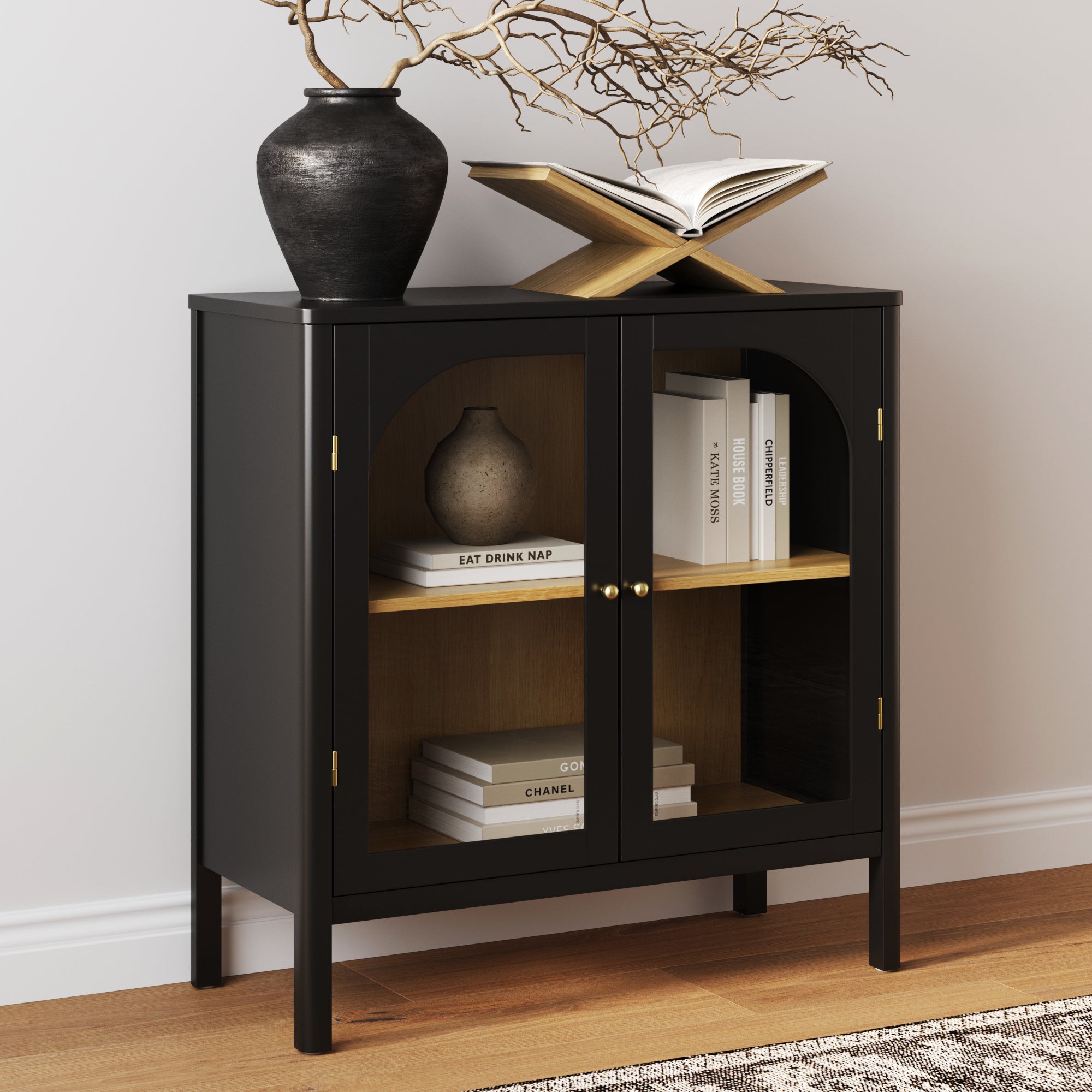 The Nathan James Wood Curio Cabinet with Glass Doors in black showcases books and decor, accented by brass hardware. On top, a black vase with bare branches and an open book on a wooden stand rest against a white wall on the wood floor.