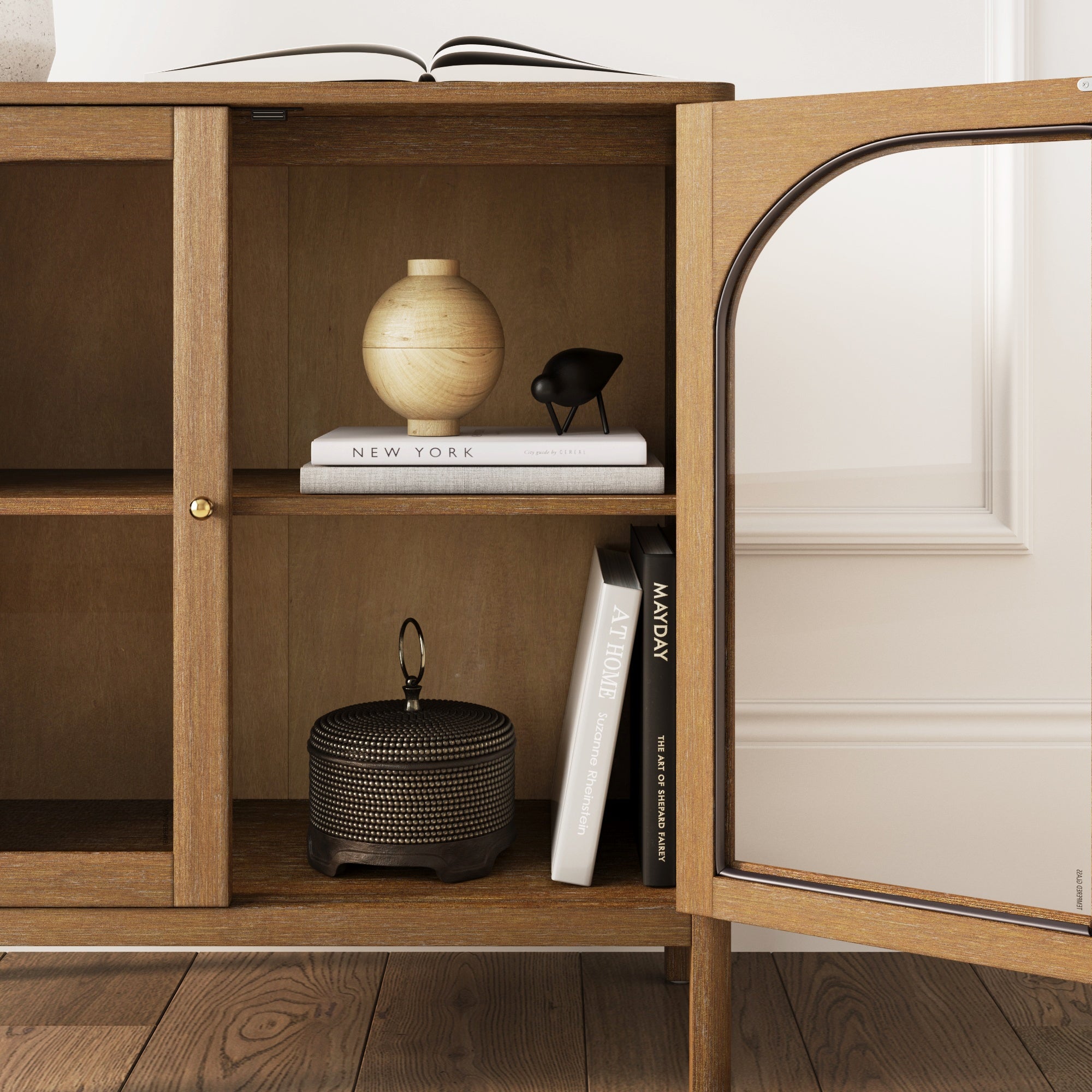 A wooden cabinet with an open glass door displays a light-colored vase, a small black bird sculpture, stacked books, and a textured decorative container on wooden shelves. The floor is dark wood and the background is a white wall.