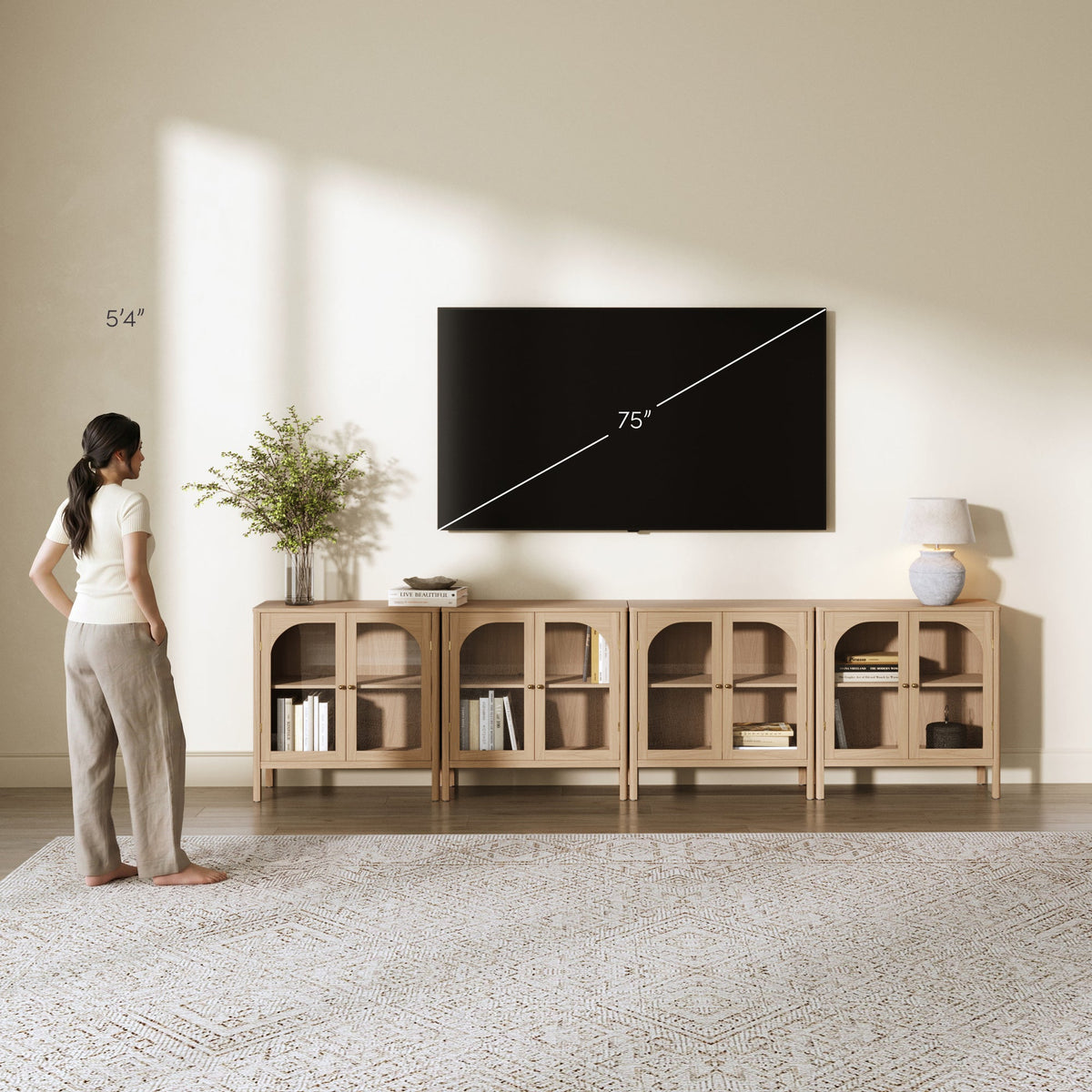 A person stands before a set of four Nathan James Large Glass Door Curio Cabinet Credenza Raw Oak units with glass-paneled doors and brass hardware, positioned under a wall-mounted 75-inch television.