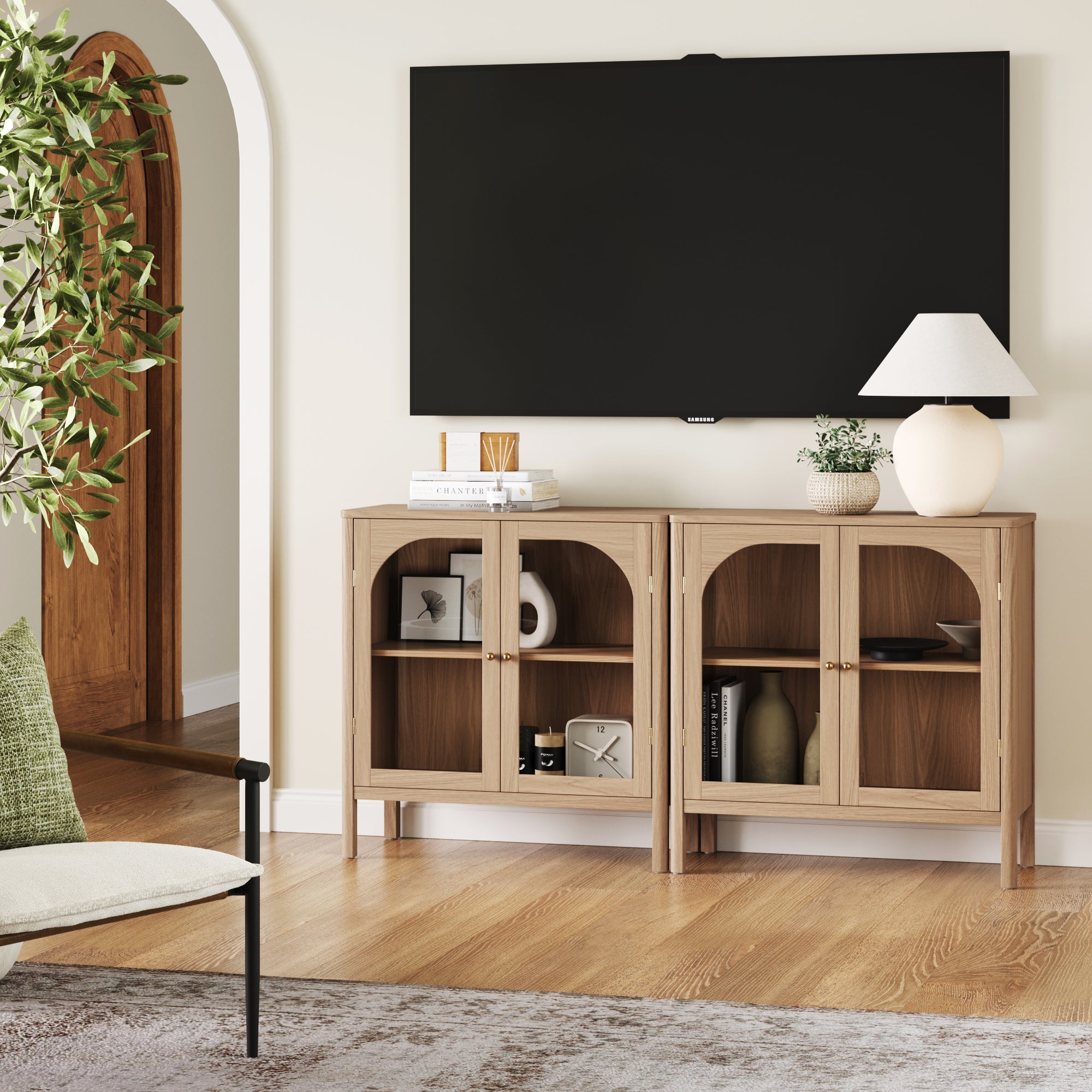 A modern living room showcases the Nathan James Curio Cabinet TV Stand Credenza with Glass Door in Raw Oak (Set of 2) beneath a large wall-mounted TV, paired with books, decor, a lamp, and a green-cushioned chair for an inviting vibe.
