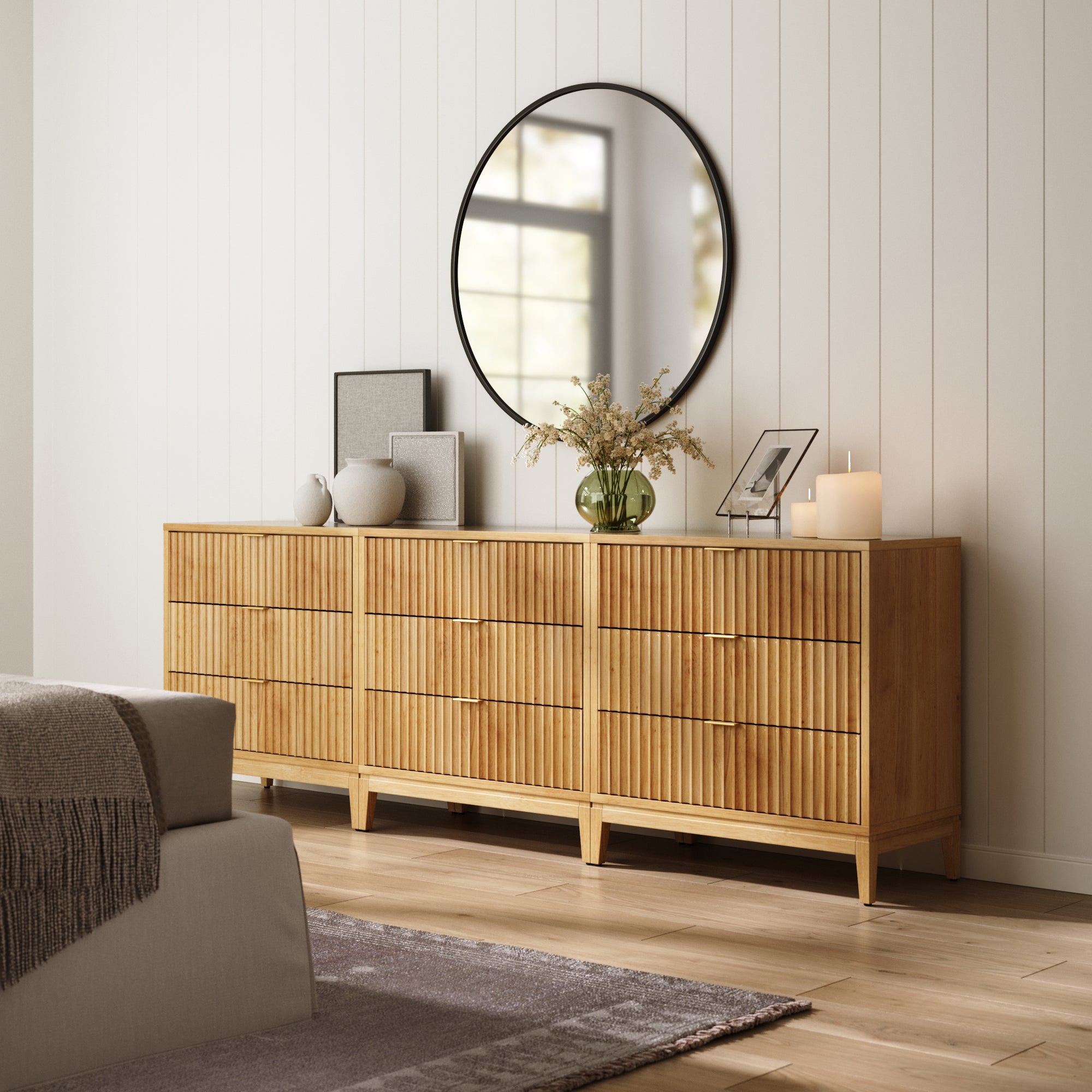The Nathan James Mid-Century Fluted 3-Drawer Dressers in Warm Pine (Set of 3) stands against a white paneled wall, adorned with a round mirror, framed art, vases with flowers, and a lit candle. A gray sofa and rug are partially visible.