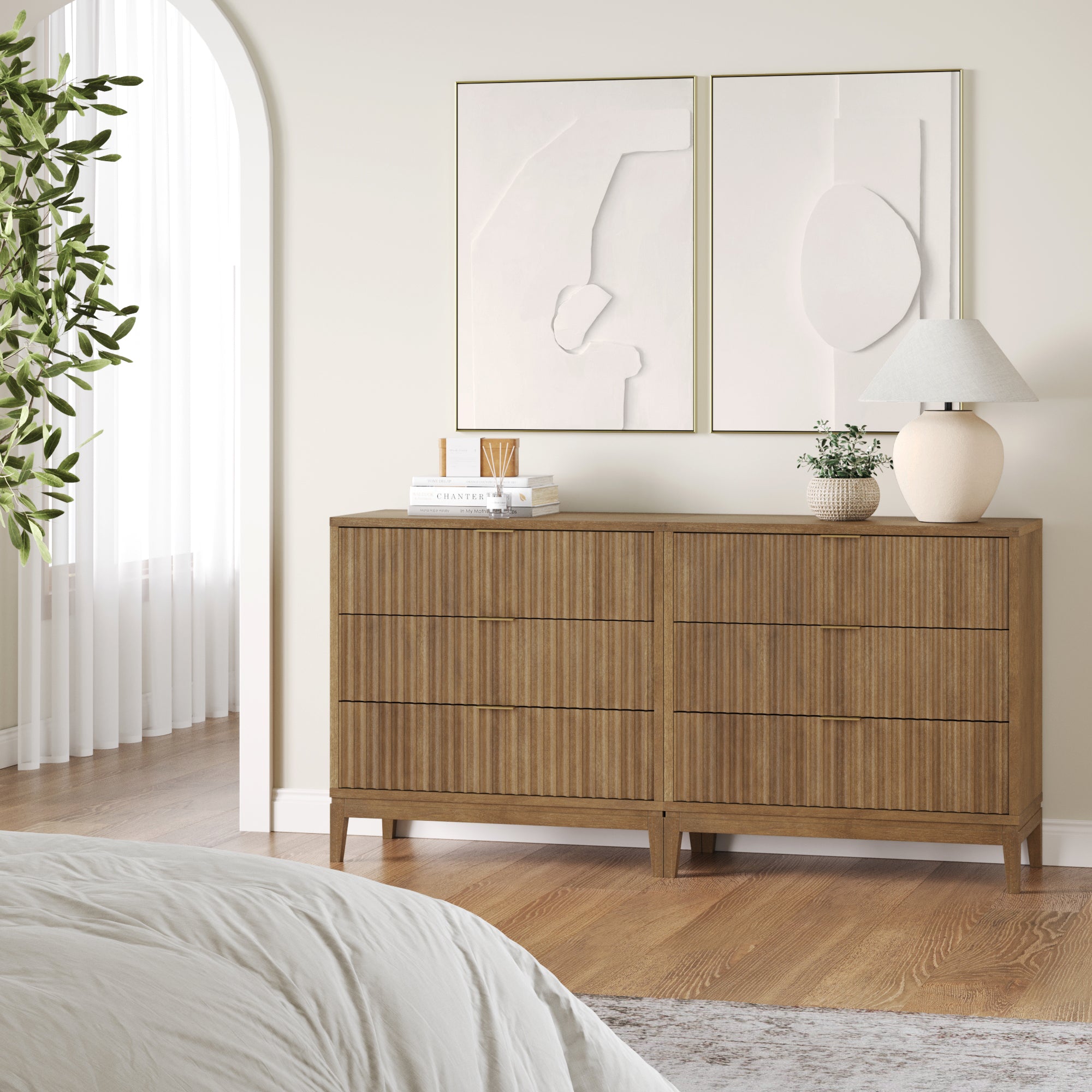 A modern bedroom featuring two Nathan James Bohemian Solid Wood 3-Drawer Fluted Dressers in light brown, minimalist abstract wall art, books, a table lamp, a potted plant, and a bed with white bedding in the foreground.