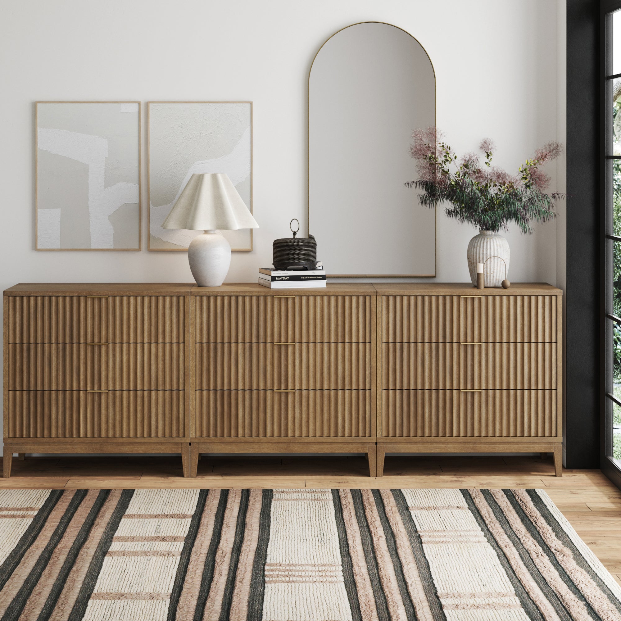 A modern wooden sideboard with vertical grooves stands against a white wall, topped with abstract art, a lamp, a round mirror, decorative items, and a vase with foliage. A striped rug covers the wooden floor.
