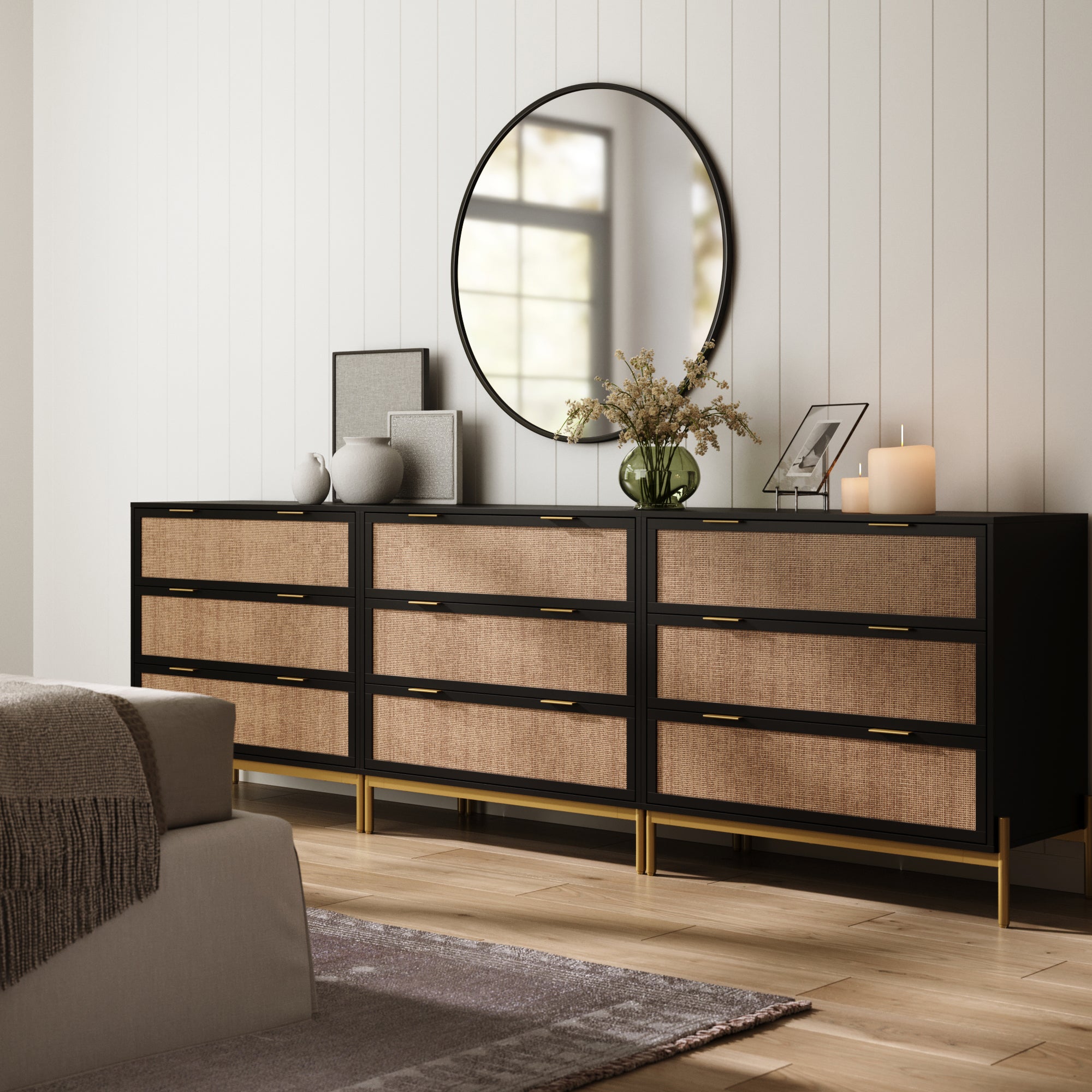 The Nathan James Rattan & Wood 3-Drawer Dressers in black (set of 3) feature cane fronts and gold handles. Topped with decor, flowers, art, and candles, they sit below a large round mirror reflecting light from a bright window.