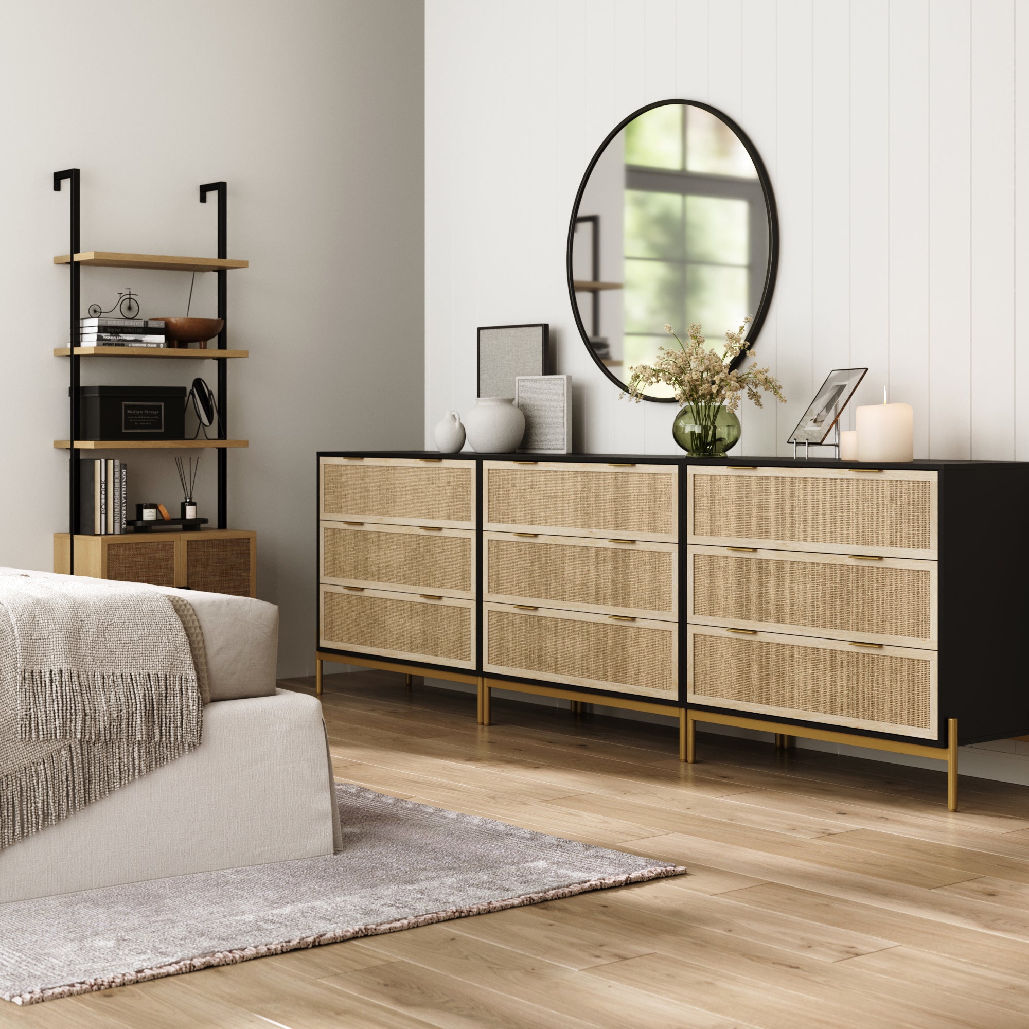 A modern bedroom with a beige bed, gray rug, and three black Nathan James Rattan & Wood 3-Drawer Dressers. Above, a round mirror hangs, while shelves display decorative items and books, all on a light wood floor.
