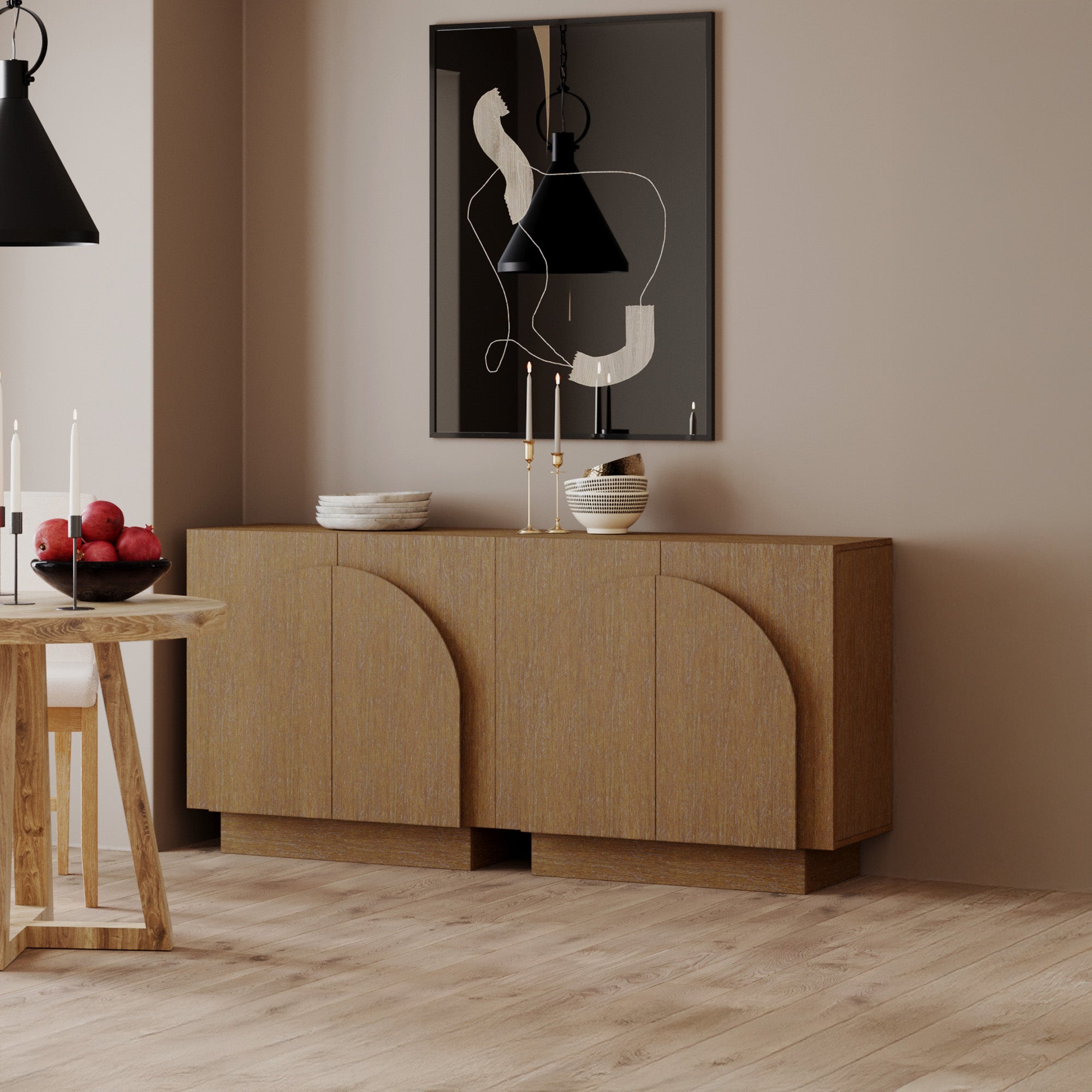 A modern dining room with light wood flooring, a wooden sideboard with arched details, abstract wall art, black pendant lights, and a round table topped with apples and candles. Neutral color scheme throughout.
