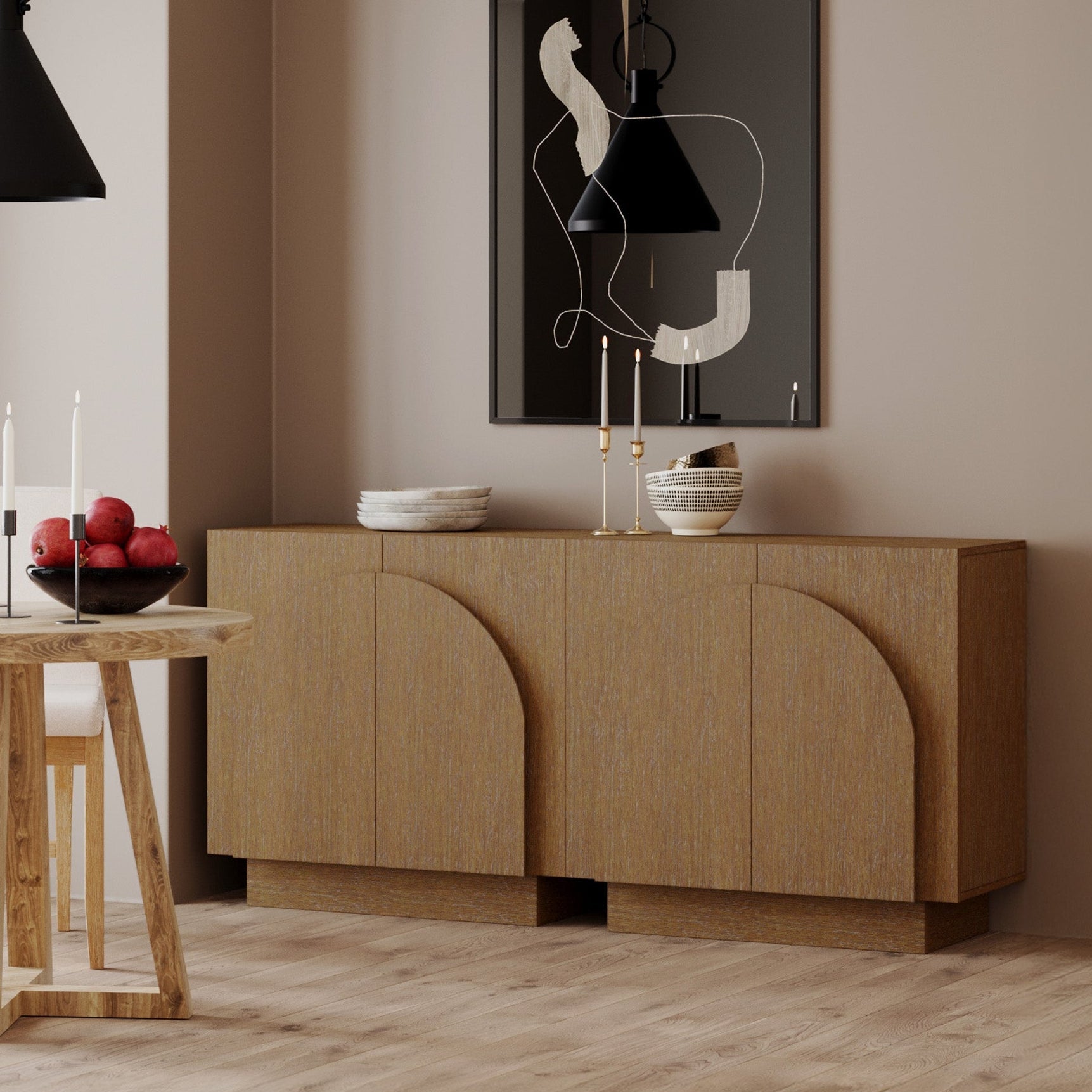 A modern dining room with light wood floors, a wooden sideboard with curved designs, a table with candles and a bowl of red apples, black pendant lights, and abstract wall art in a black frame.