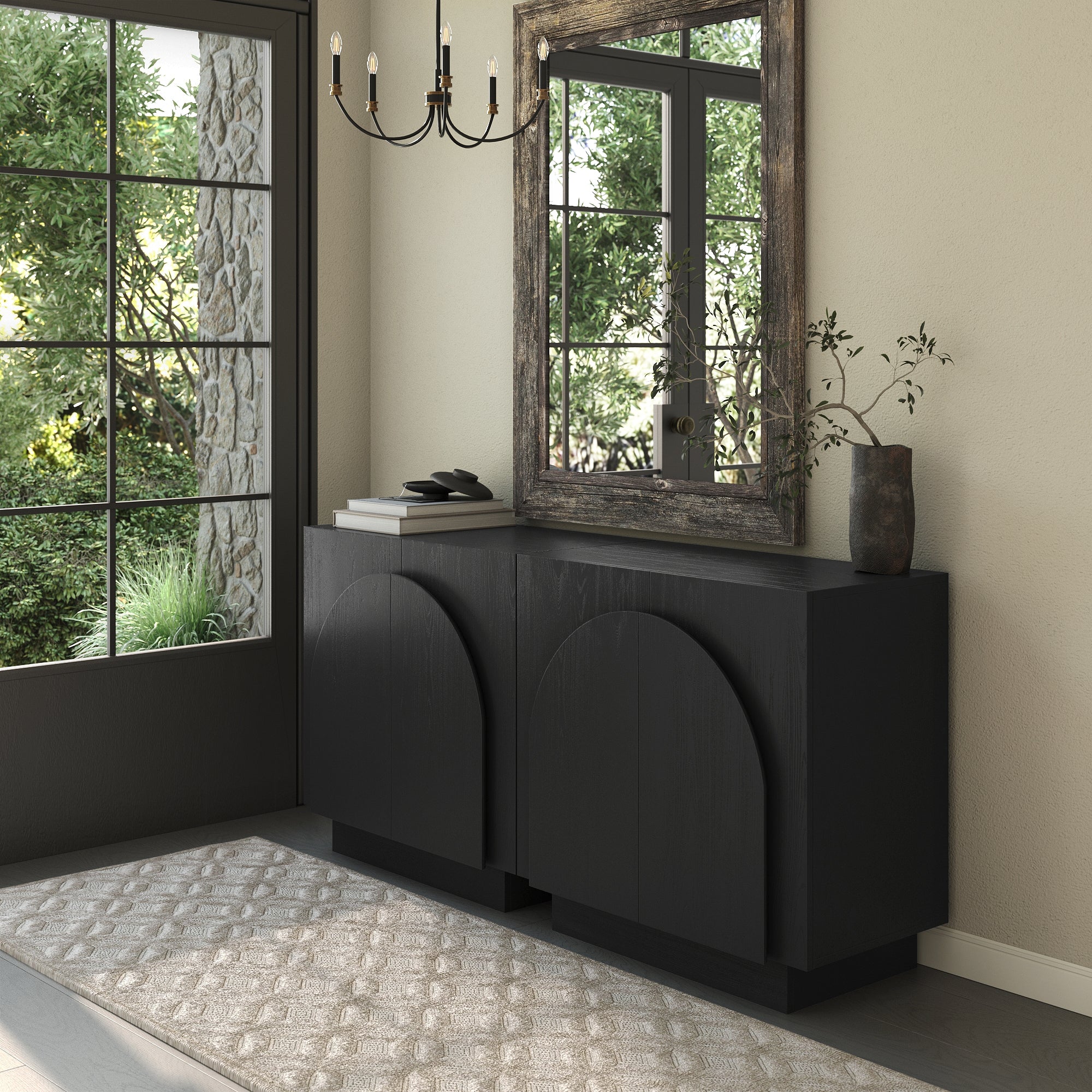 A modern entryway with a large window, black sideboard with curved details, a decorative vase with green branches, a stack of books, a rustic framed mirror, chandelier, and a patterned beige rug.