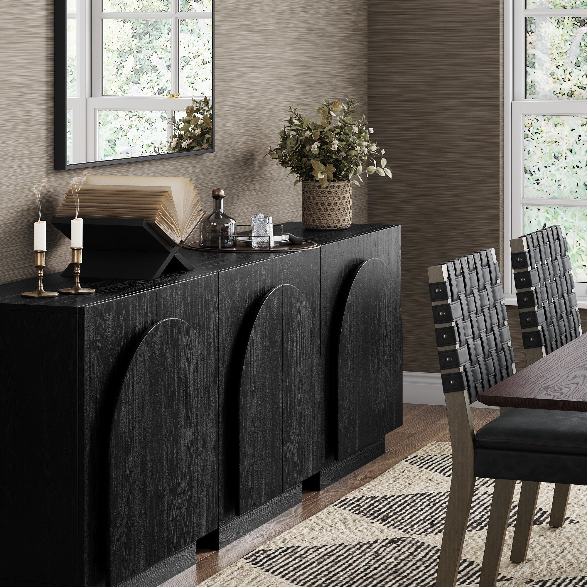 A modern dining room with a black sideboard, two candles, an open book, a plant in a decorative pot, a large mirror above, and a wooden dining table with woven black chairs on a patterned rug.