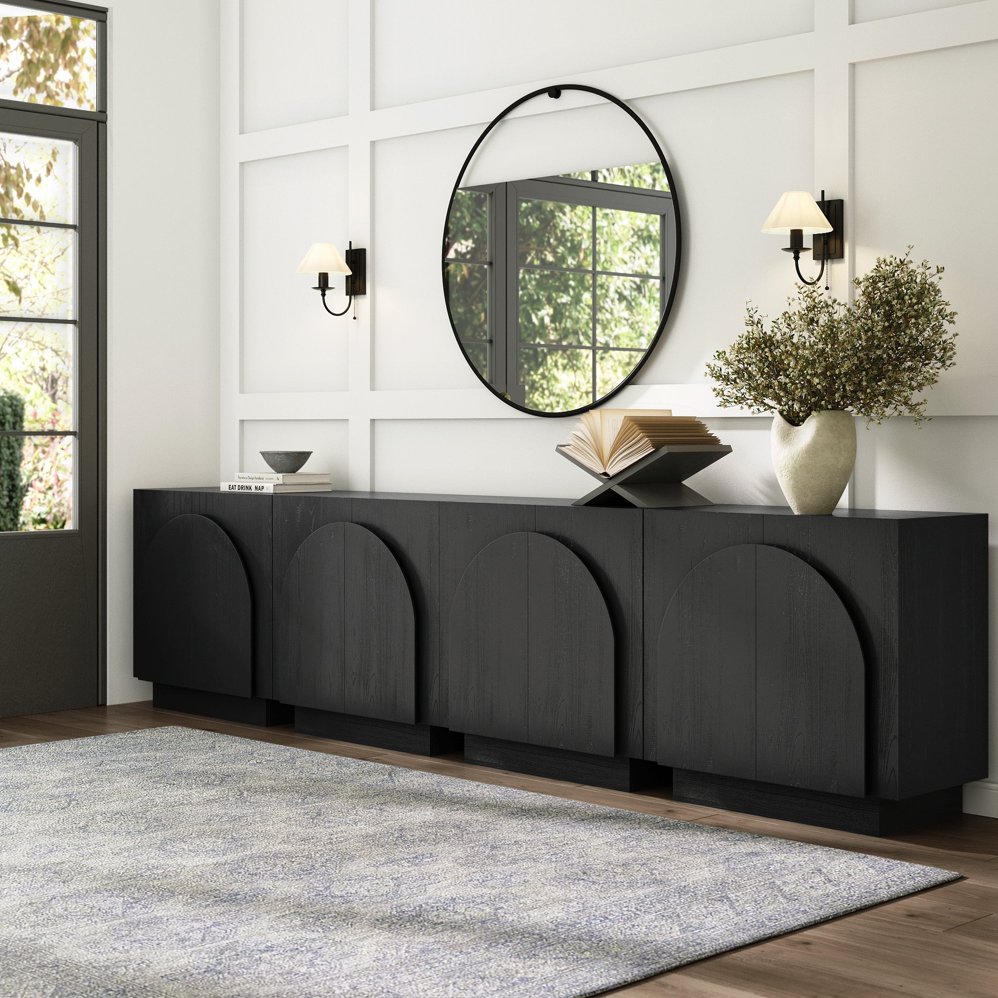 A modern entryway features a black sideboard with arched detailing, a large round mirror above it, wall sconces on each side, a vase with branches, an open book, and a gray rug on the floor.