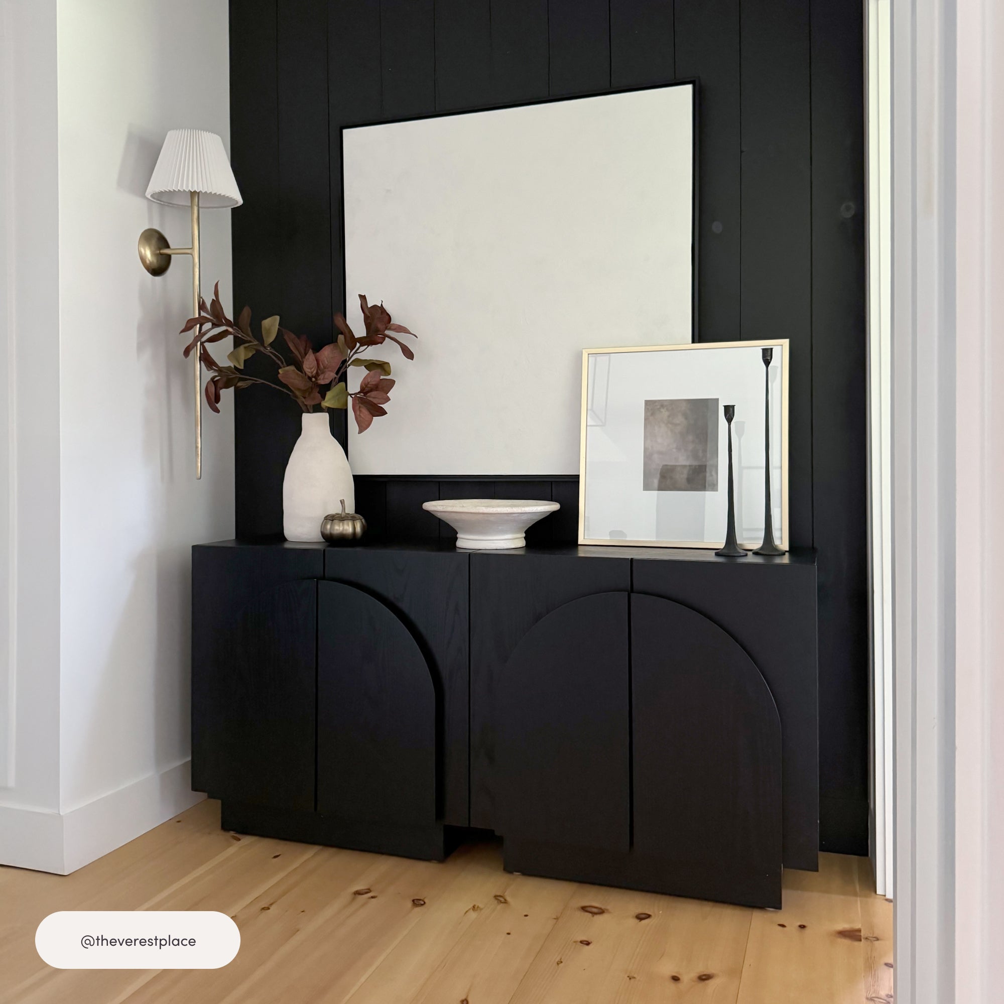 A modern entryway with a black cabinet, large white vase with branches, white bowl, two framed abstract artworks, and a wall-mounted white sconce, all set against a black paneled wall and light wood floor.