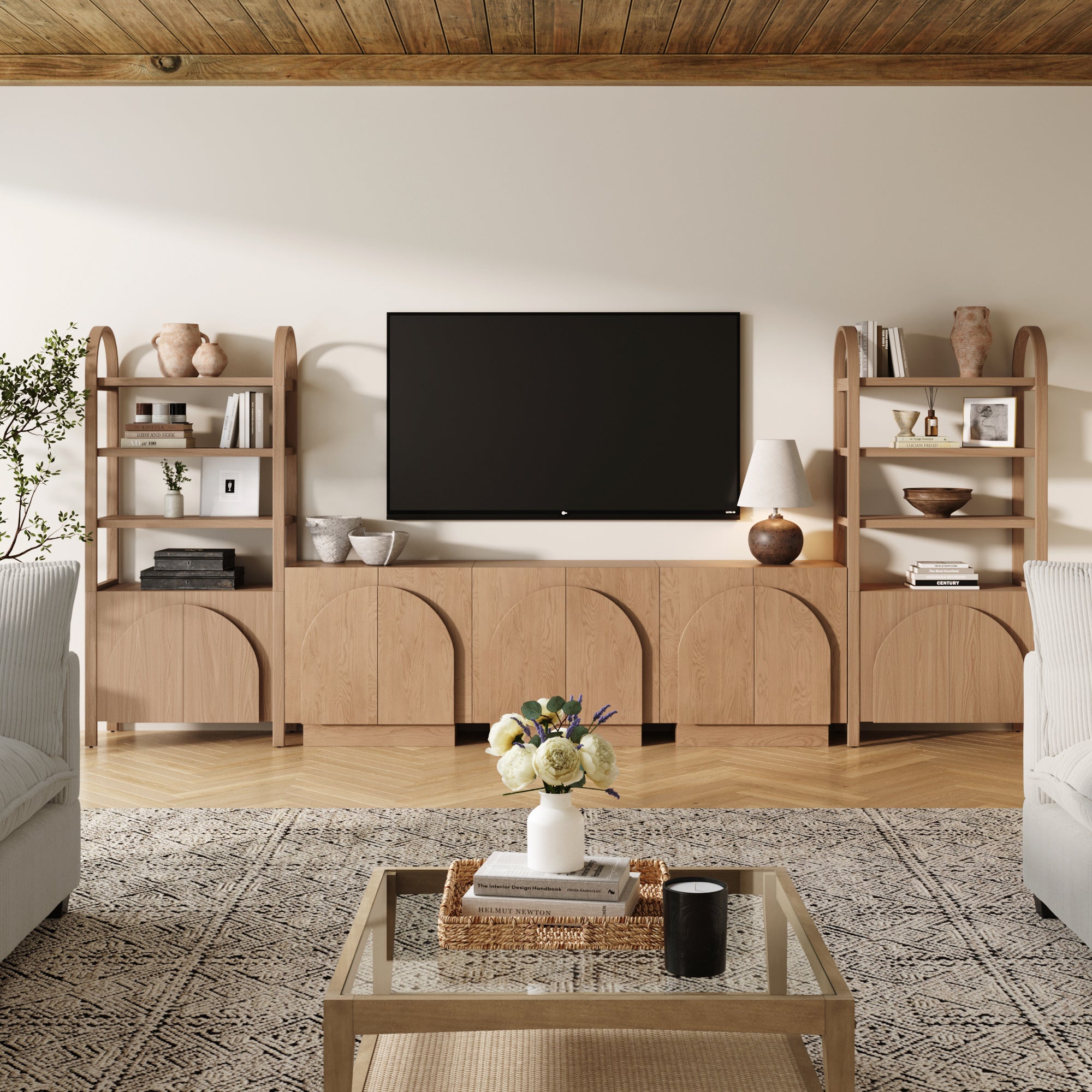A modern living room features a large TV on the Nathan James Mid-Century Modern Arched Wood Entertainment Center (3 Cabinets), flanked by matching shelves. A glass coffee table with books and flowers sits on a patterned rug between two sofas.