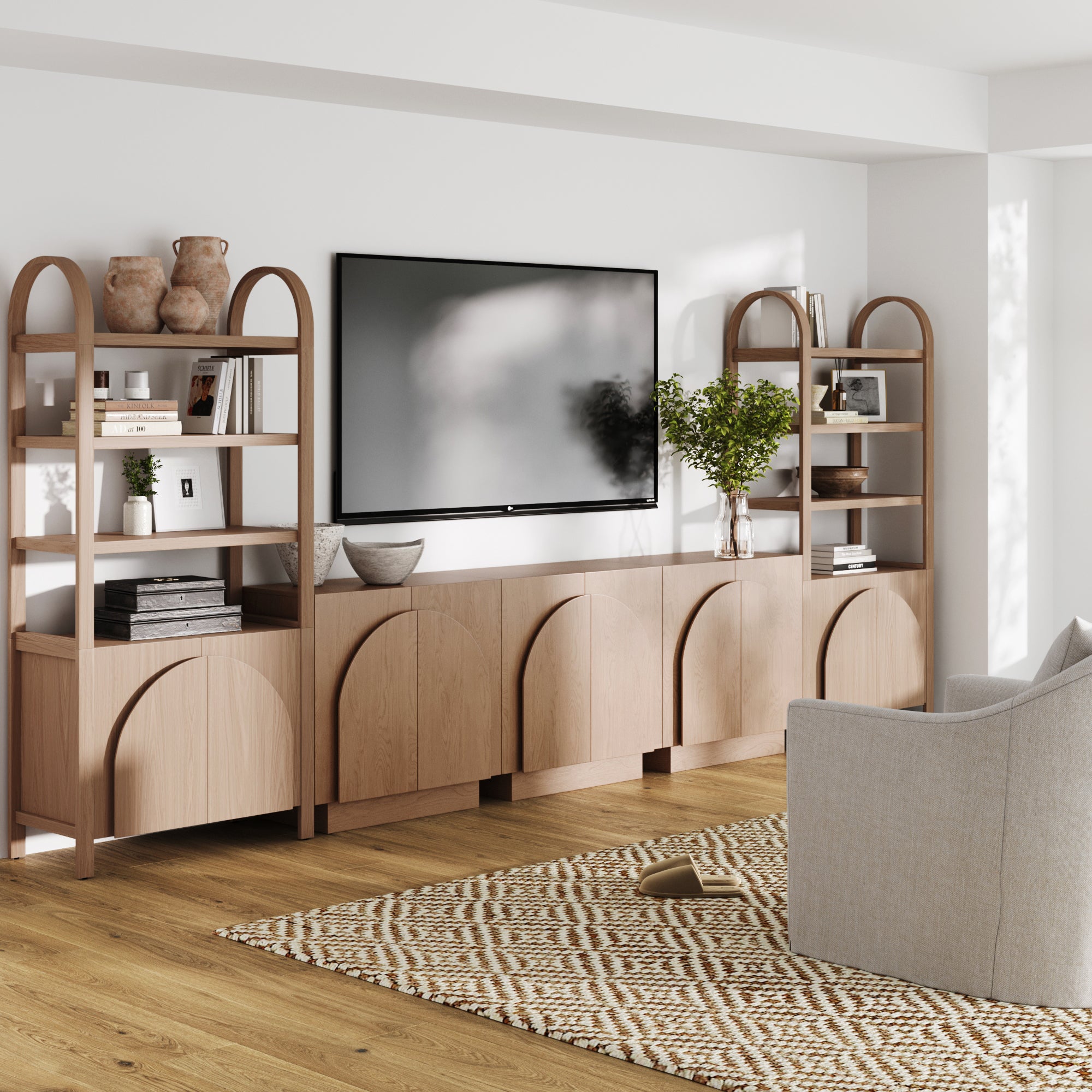 A modern living room showcases a wall-mounted TV above the Nathan James Mid-Century Modern Arched Wood Entertainment Center (3 Cabinets), with light wood cabinets, open shelves, books, vases, plants, a patterned rug, and a light gray armchair on wood floors.