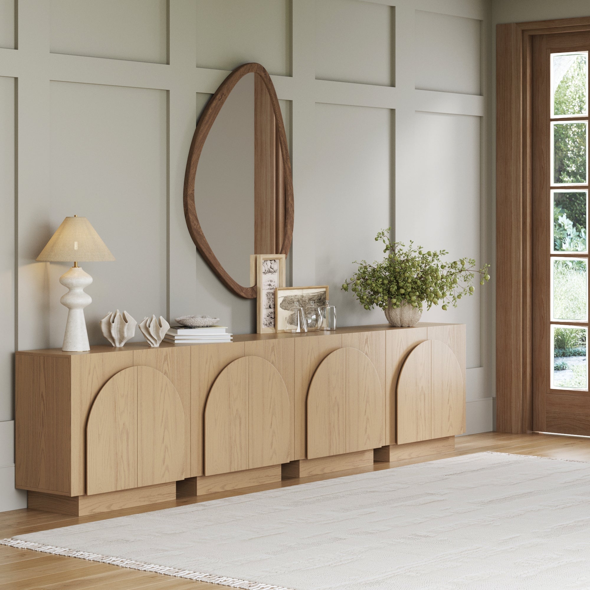 A modern living room features the Nathan James Wood Mid-Century Arched Storage Cabinet in Gray Oak (Set of 4) with arched doors, an oval mirror above, decor accents, a lamp, plants, and large windows bringing in natural light for a mid-century vibe.