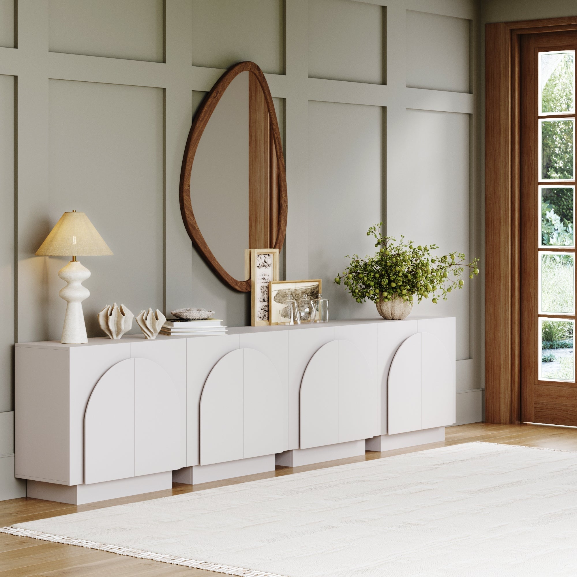 A modern living room with a light gray paneled wall, white arched sideboard, table lamp, books, decorative objects, green plant, oval wooden mirror, and a large window letting in natural light.